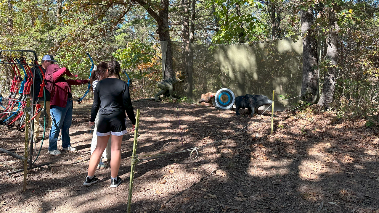 People watching a small dinosaur-themed display with life-sized models of a T-Rex, dinosaur figures, a bear, and a target in a wooded area.