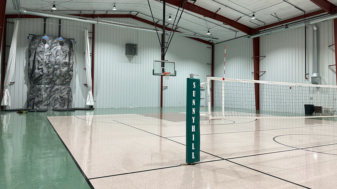 Indoor volleyball court with a net and a volleyball pole, a rock climbing wall on the left side, and a basketball hoop on the back wall.