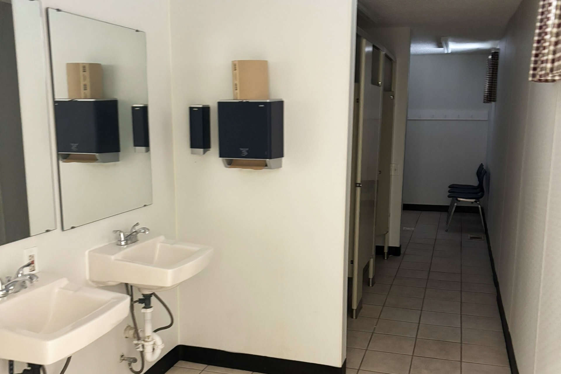Public restroom with two white sinks, mirrors, black paper towel dispensers, and a row of chairs in the hallway.