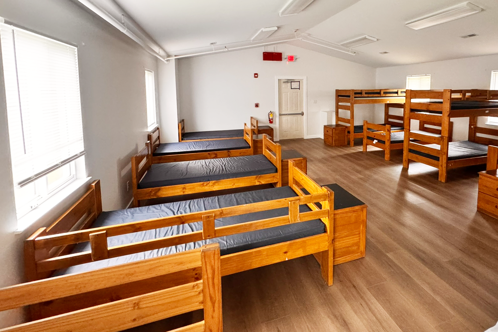 Dormitory room with multiple wooden bunk beds arranged along the walls, black mattresses on each bunk, and hardwood floors. There are windows with blinds, a door at the far end, and emergency equipment on the walls.