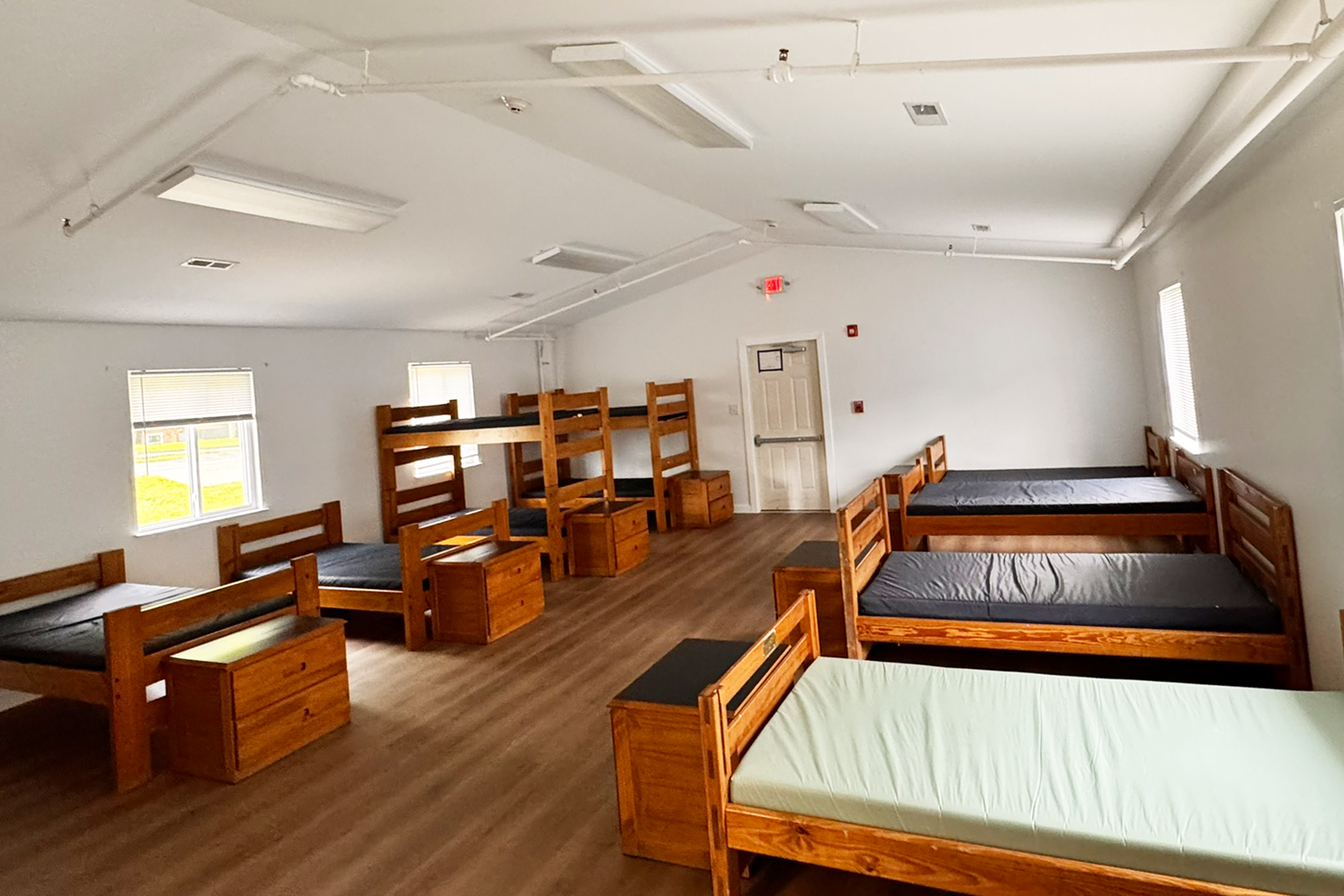 Room with multiple wooden beds arranged along the walls, some with mattresses, in a dormitory or hostel setting with hardwood floors and windows with blinds.