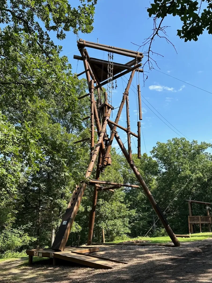 A tall wooden jungle gym or climbing structure in a wooded outdoor setting, with a clear blue sky overhead.