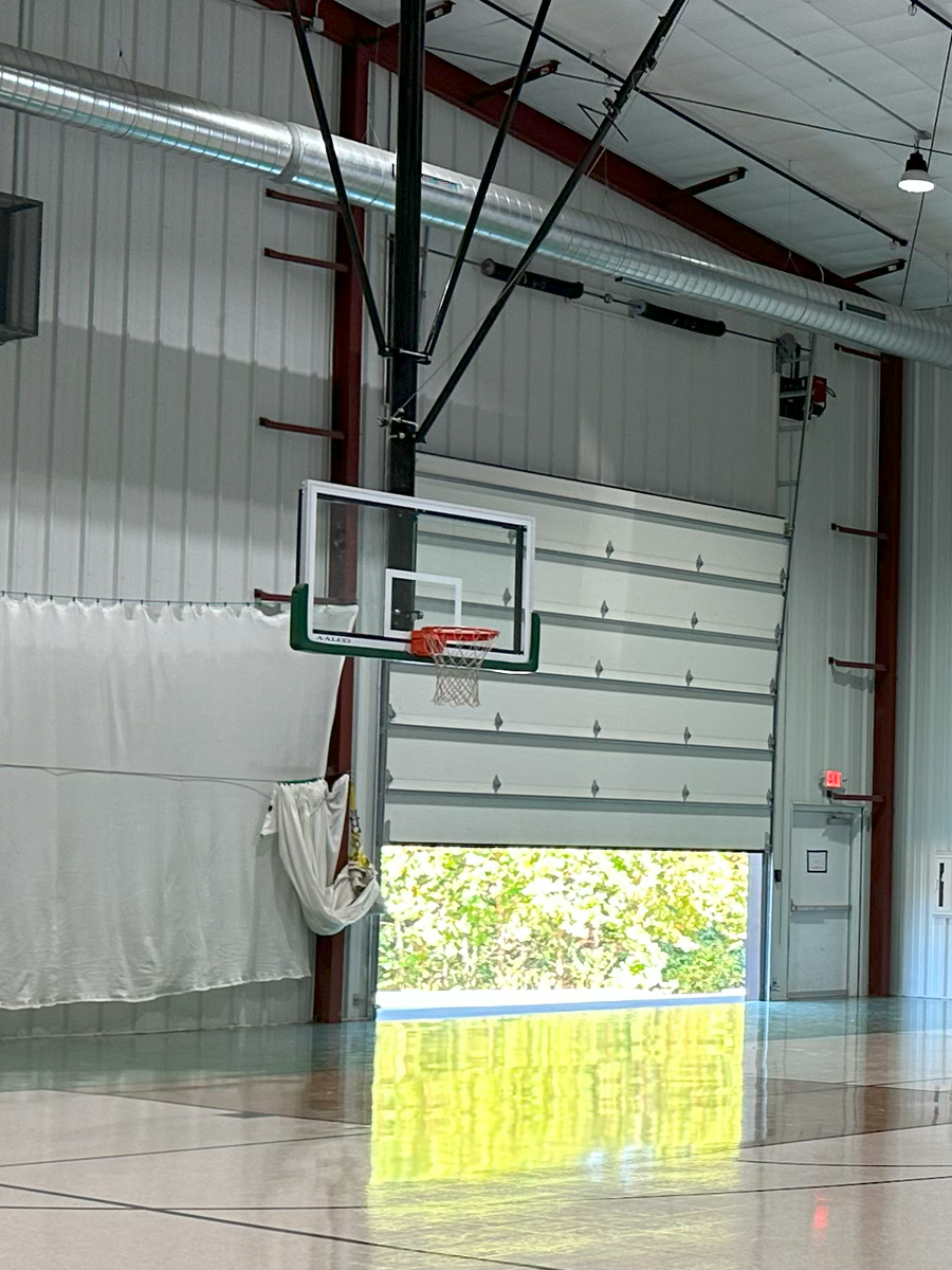 An indoor basketball court with a basketball hoop, a large garage-style door open to outside greenery, and overhead lighting and ventilation.