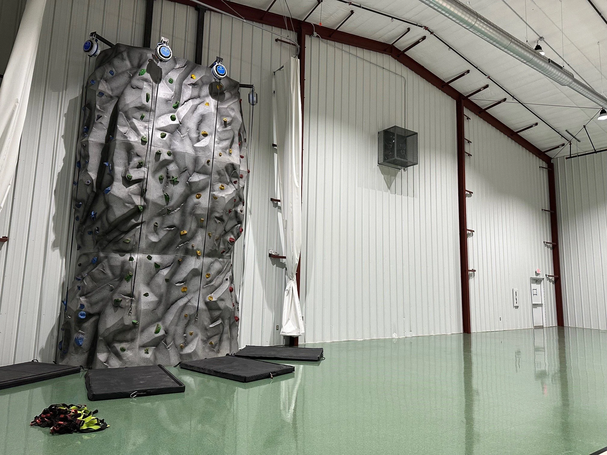 Indoor rock climbing wall with multiple climbing holds, mats on the floor, and a white curtain near the wall.