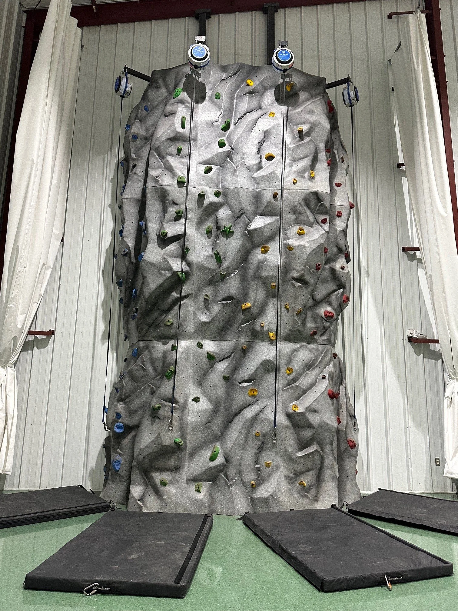 Indoor rock climbing wall with colorful holds, safety mats below, and ropes attached for climbing.