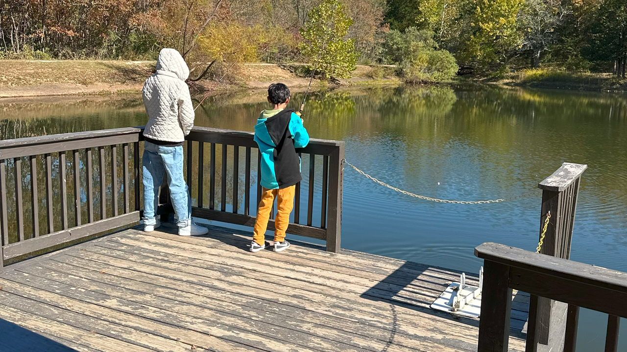 Two children, one in a beige quilted hoodie and jeans and the other in a black and turquoise jacket with yellow pants, stand on a wooden dock fishing in a calm lake surrounded by trees with autumn foliage.