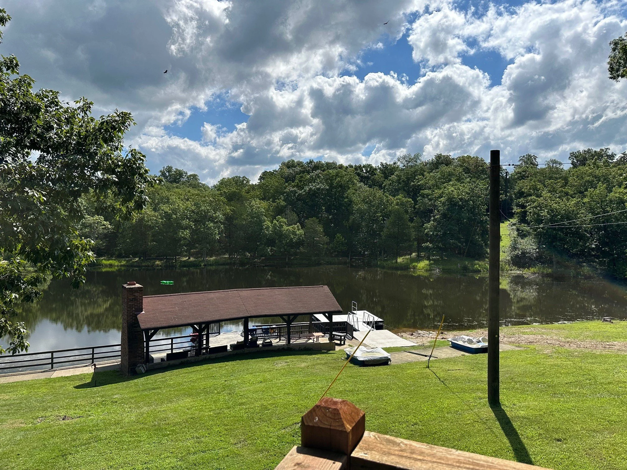 A lakeside scene with a grassy lawn, a wooden pavilion with a brick chimney, boats on the shore, and a calm lake surrounded by trees under a partly cloudy sky.