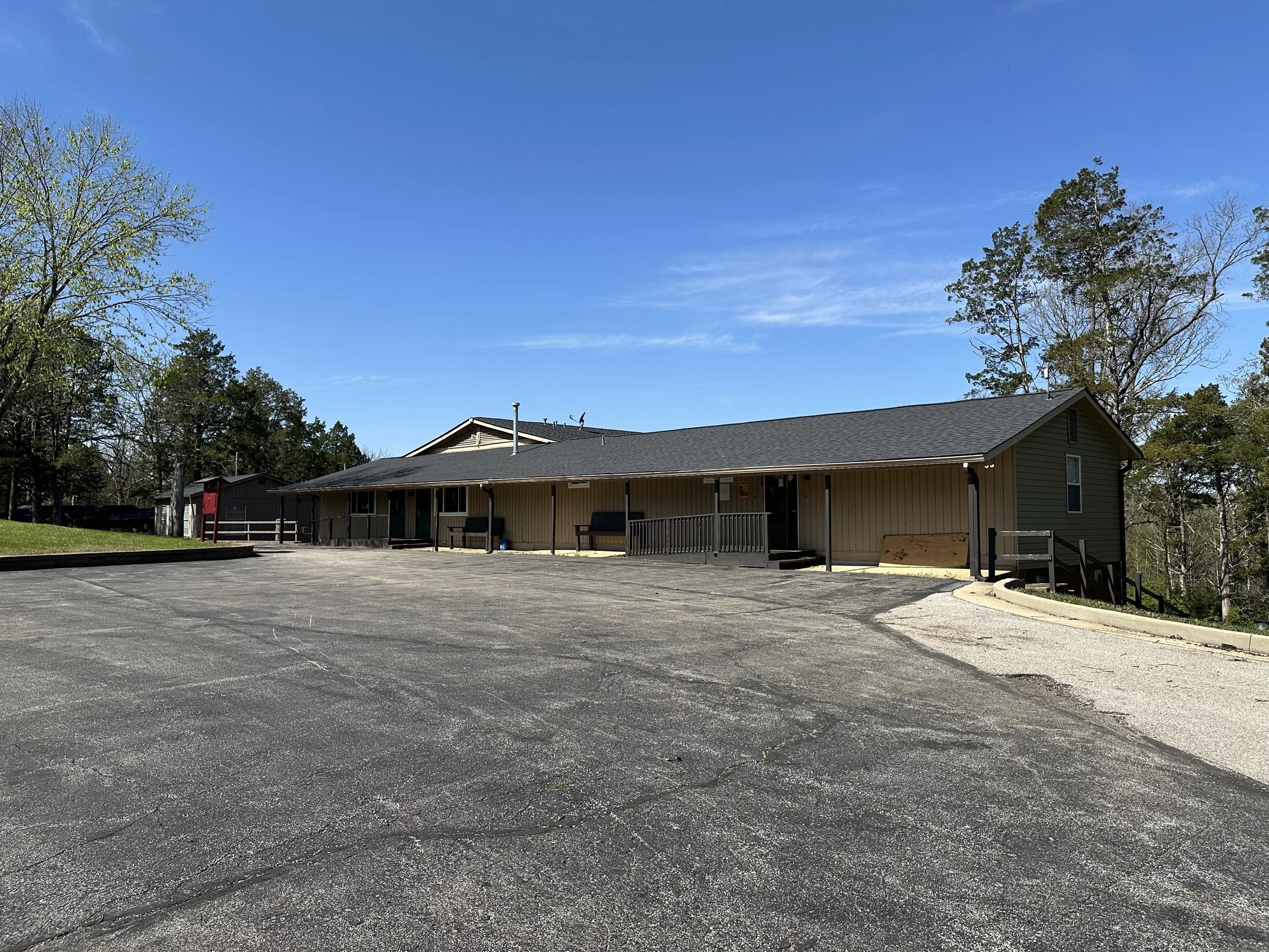 Single-story building with a gray roof, beige siding, and a large parking lot in front, surrounded by trees under a blue sky.