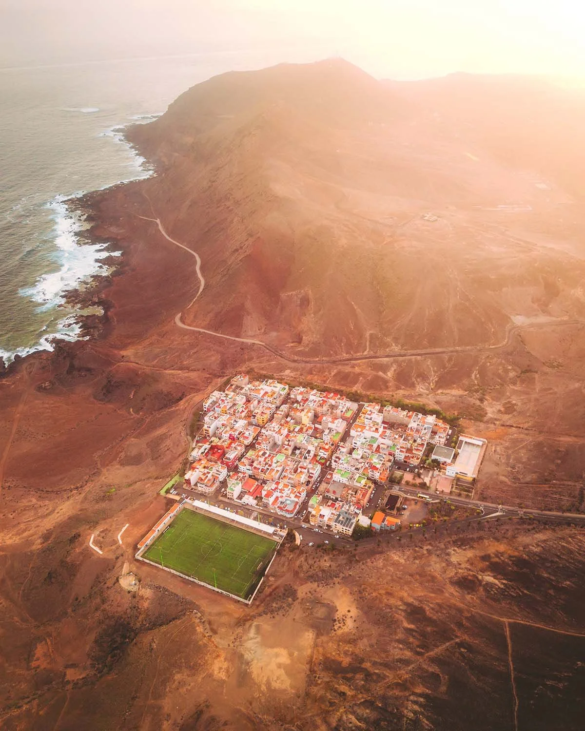 Vue aérienne d'un village côtier canarien niché au pied d'une montagne rouge, avec un terrain de football vert vif bordant l'océan.