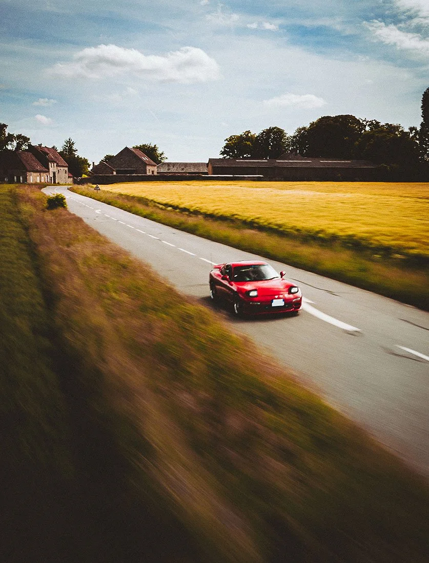 Prise de vue dynamique (rolling shot) d'une Mazda RX-7 rouge roulant sur une route de campagne, capturée par drone ou voiture suiveuse.