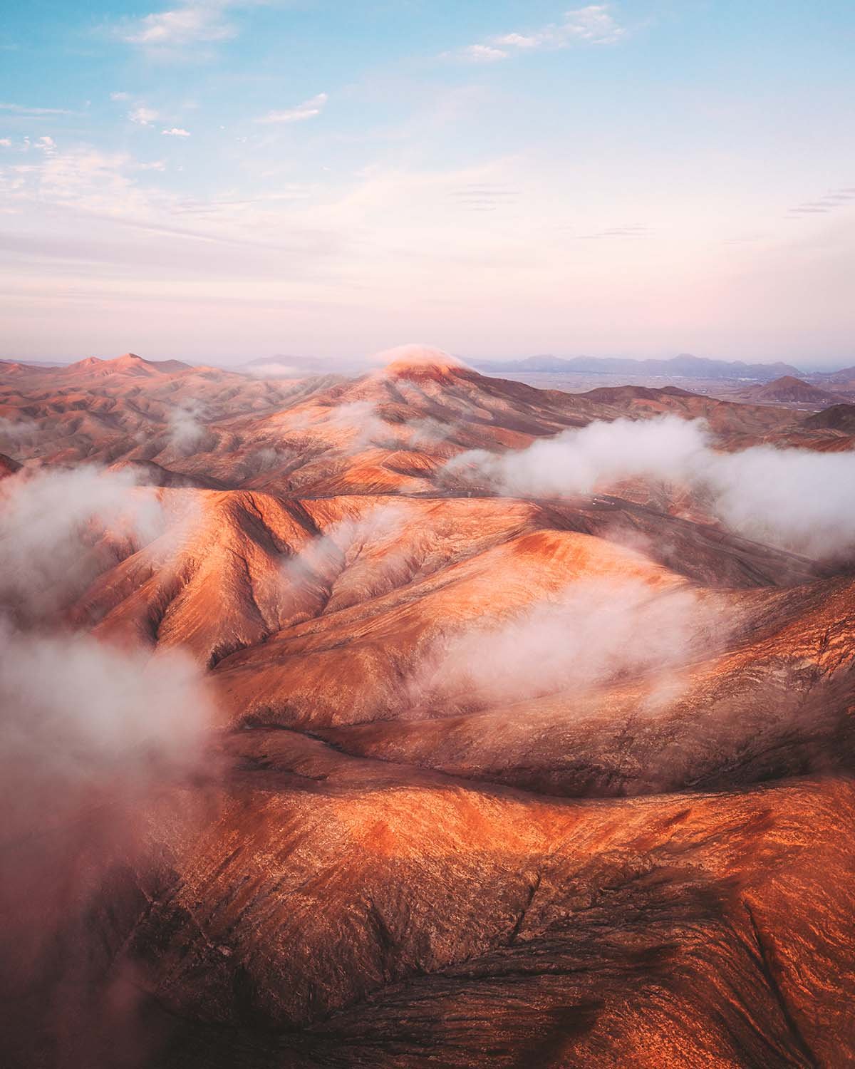 Paysage onirique de montagnes aux teintes pastel et ocre enveloppées par des nuages bas, typique de l'intérieur des terres à Fuerteventura ou Lanzarote.