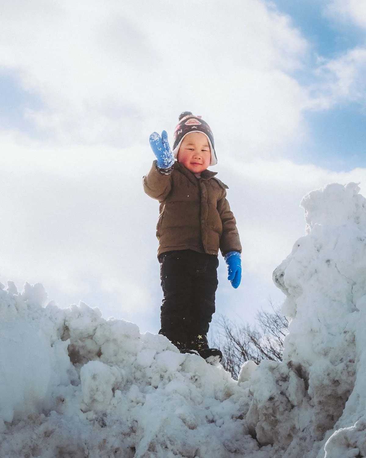 Portrait lifestyle spontané d'un enfant en tenue d'hiver saluant depuis le sommet d'un impressionnant mur de neige au Japon. Photographie de voyage en famille.