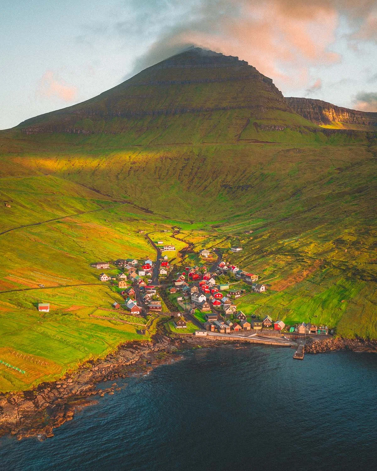Vue verticale (top-down) ou plongeante sur un village de pêcheurs typique aux maisons colorées, encaissé dans une vallée verte aux Îles Féroé.
