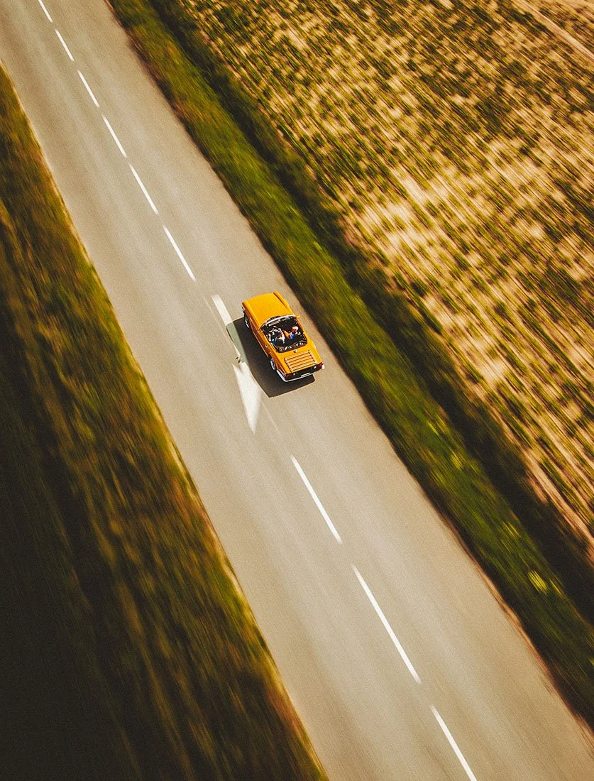 Vue aérienne zénithale d'une voiture de collection décapotable jaune roulant à travers champs. Une image évoquant l'été et la liberté.