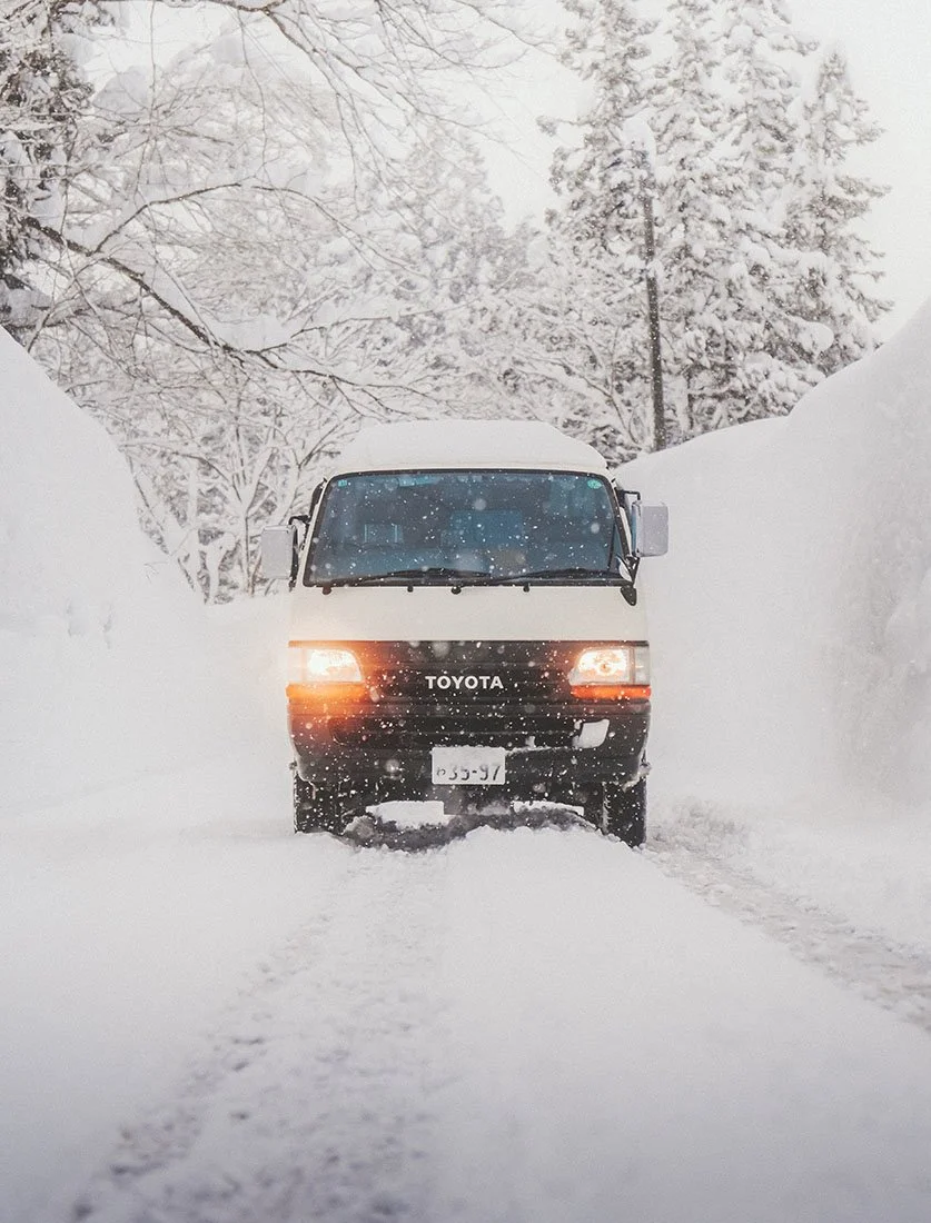 Van circulant entre d'impressionnants murs de neige lors d'un roadtrip hivernal au Japon. Photographie d'aventure et de paysage extrême.