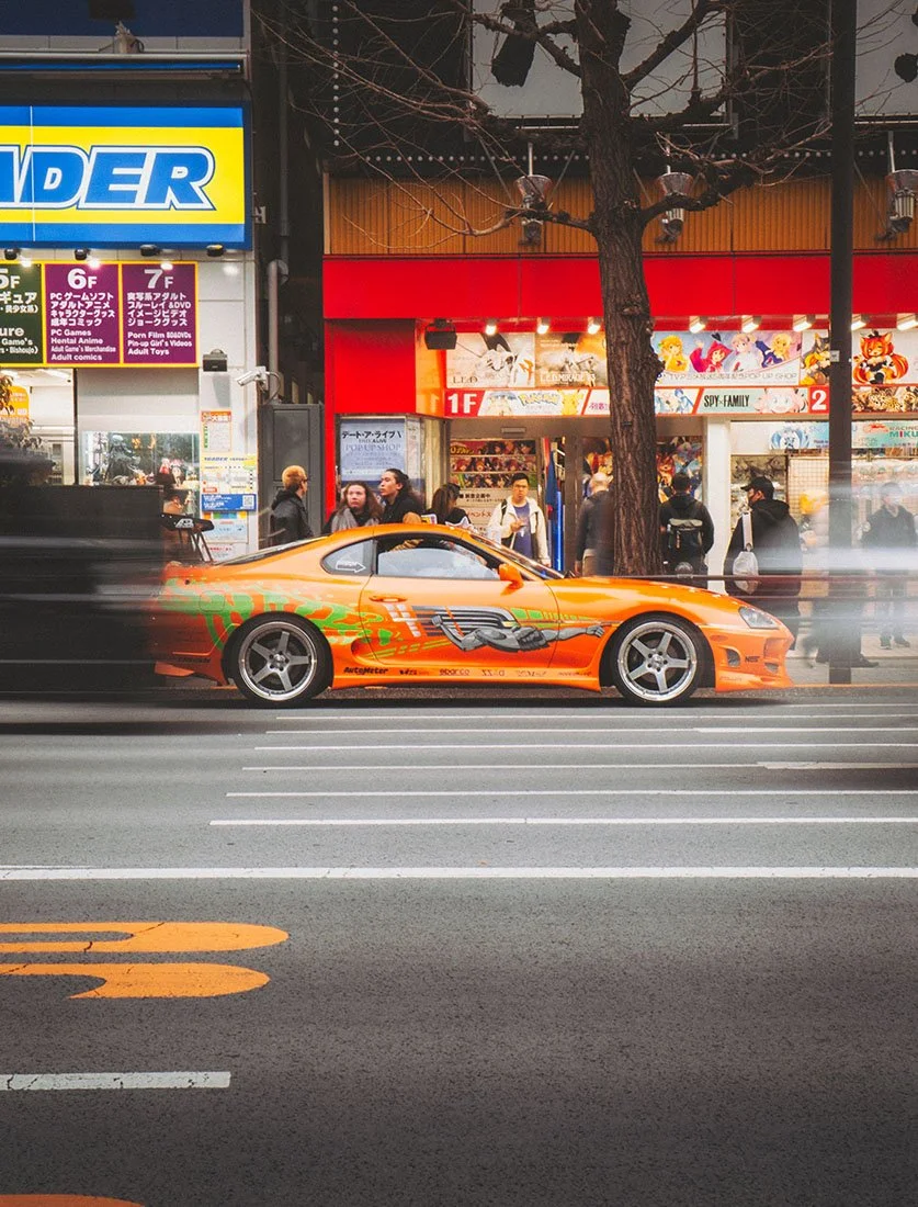 Toyota Supra orange (style Fast & Furious) circulant dans les rues animées d'Akihabara à Tokyo. Photographie automobile célébrant la culture JDM et urbaine.