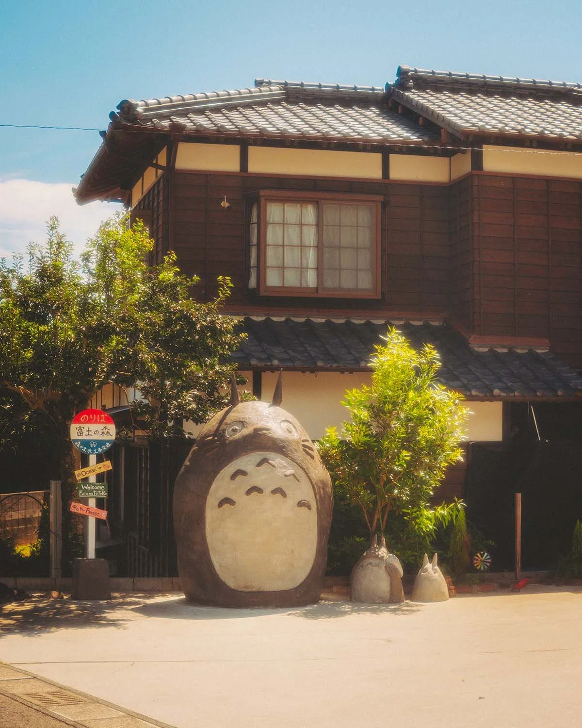 Reproduction grandeur nature de Totoro à un arrêt de bus rural au Japon. Hommage à l'animation du Studio Ghibli devant une maison traditionnelle en bois.