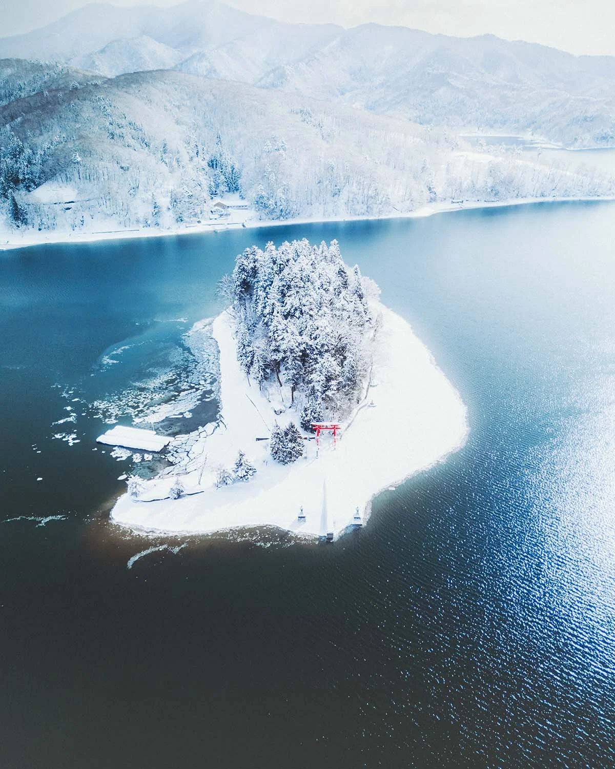 Vue aérienne féerique de l'île de Bentenjima et son torii rouge, isolés au milieu des eaux bleu sombre du lac Nojiri entouré de montagnes enneigées à Nagano.