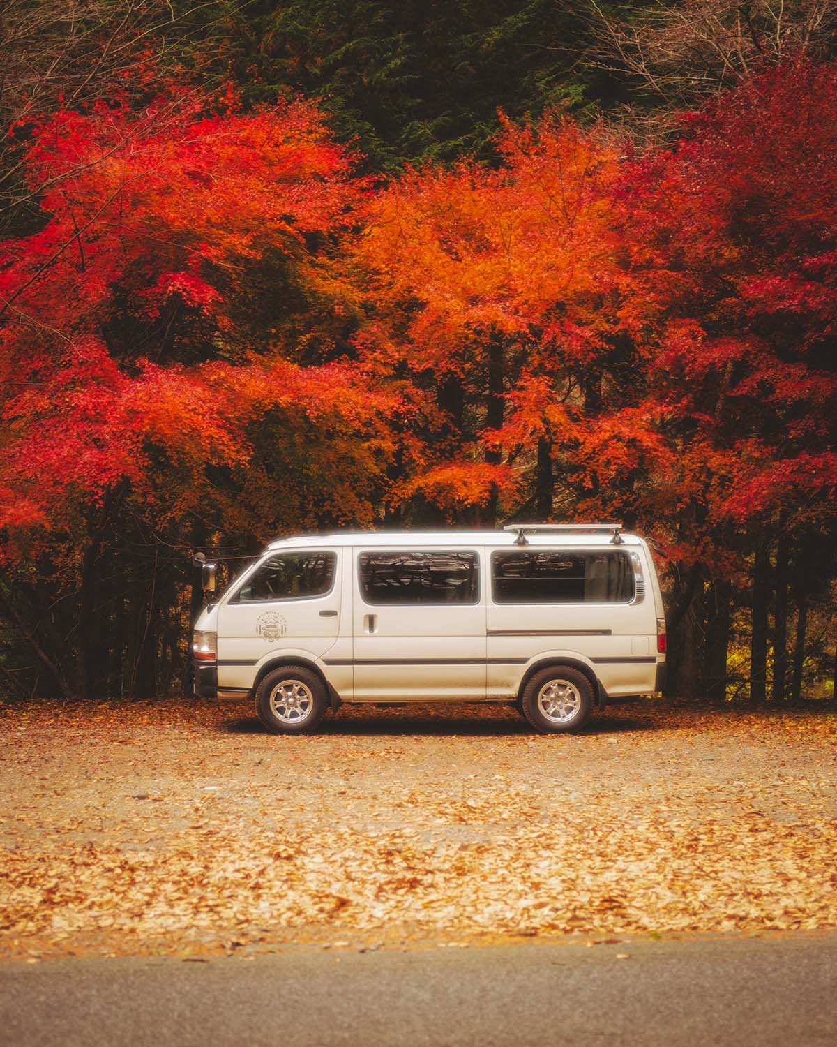 Van aménagé Toyota Hiace blanc stationné forêt automne feuilles rouges érables momiji. Roadtrip Japon vanlife camping car voyage liberté nature saisonnier véhicule japonais.