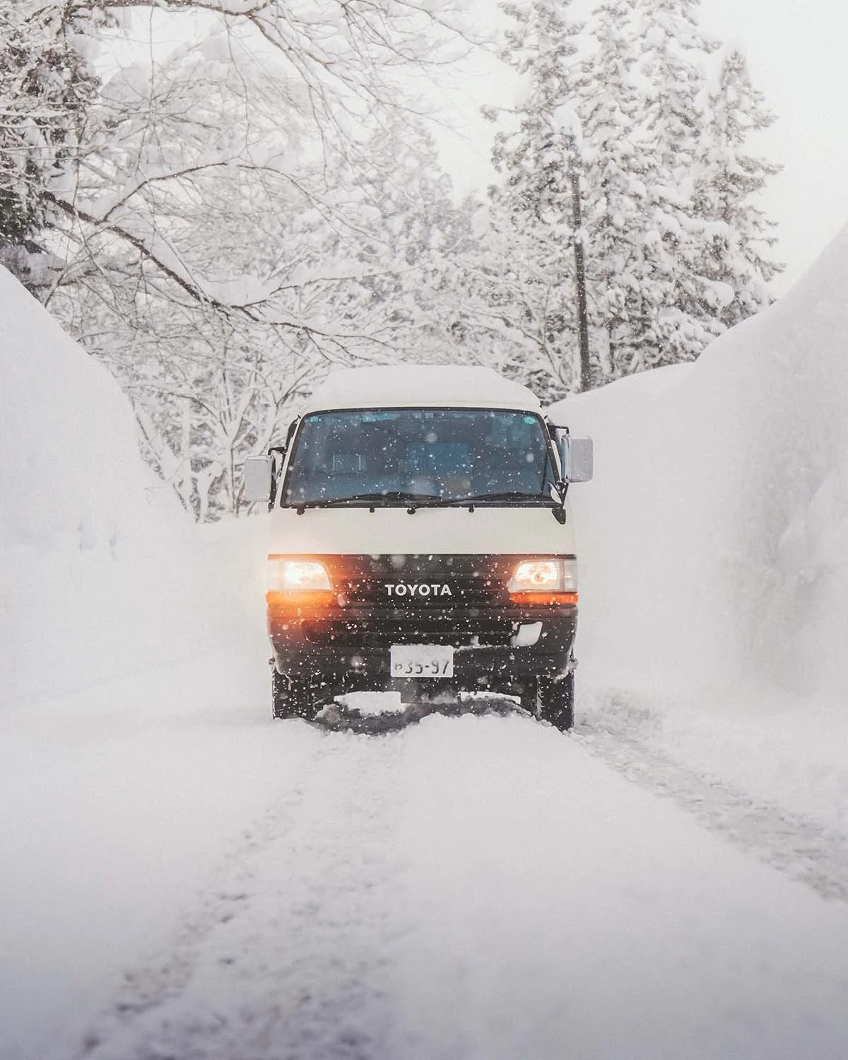 Une fourgonnette Toyota circulant difficilement entre deux murs de neige massifs sur une route de montagne japonaise. Photographie d'aventure hivernale et conditions extrêmes.