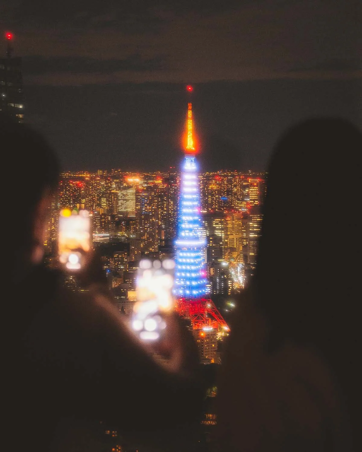 Vue nocturne illuminée Tour de Tokyo Tokyo Tower depuis observatoire Roppongi Hills avec flou artistique bokeh premier plan. Skyline ville lumière gratte-ciels architecture emblématique tourisme capitale Japon.