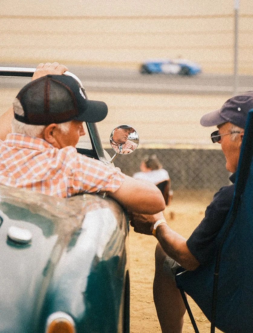 Scène de vie authentique sur circuit : passionnés échangeant autour d'une voiture de collection décapotable. Une approche lifestyle de la photographie automobile par Dimitri Weber.