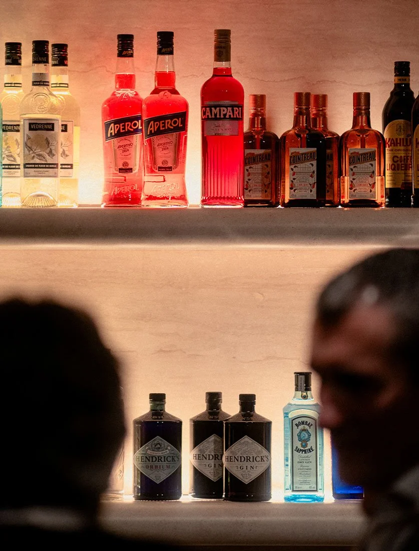 Ambiance tamisée d'un bar avec mise en valeur des spiritueux rétroéclairés et silhouettes de clients. Photographie lifestyle nocturne.