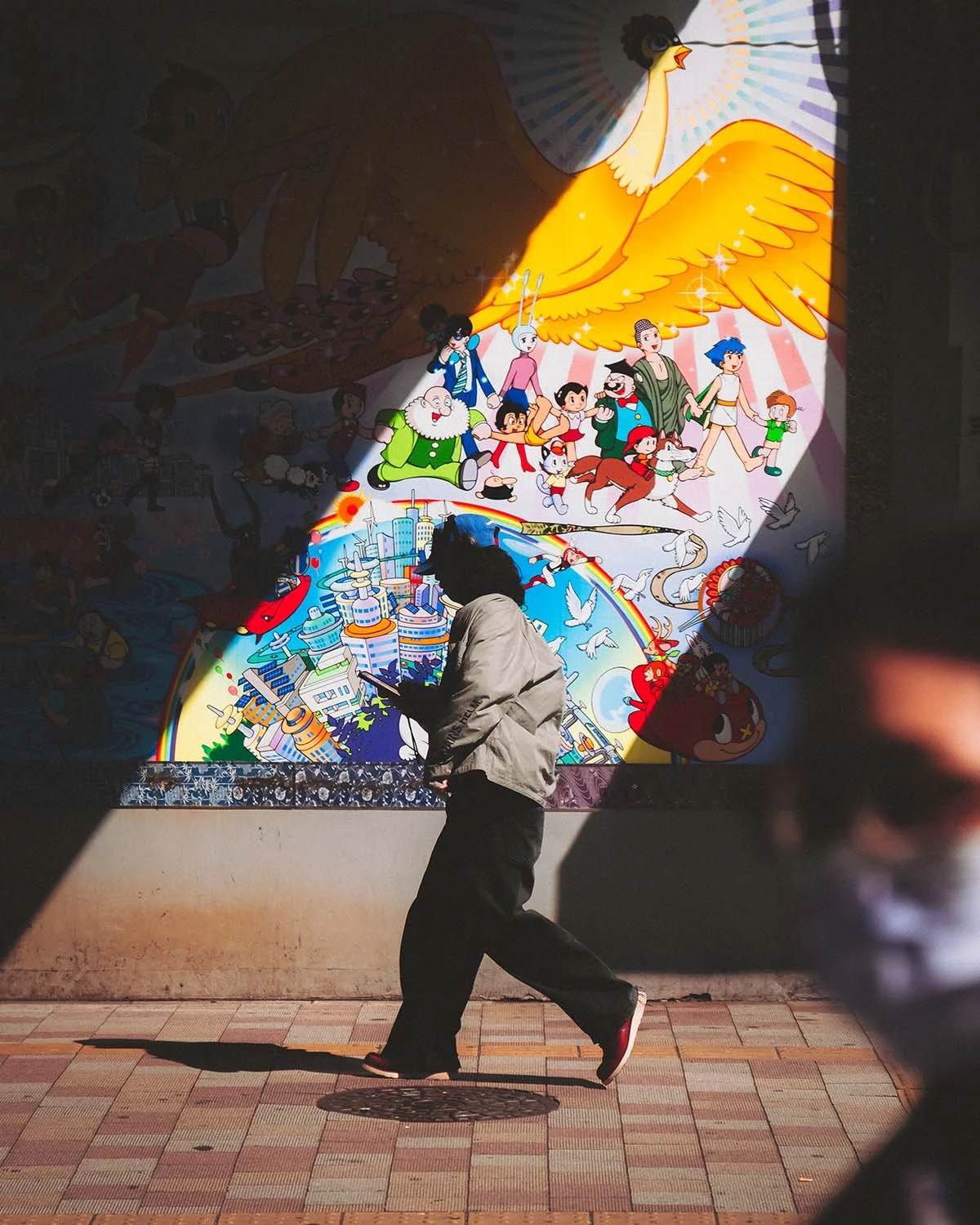 Jeu d'ombre et de lumière : un passant marche devant une fresque murale colorée représentant des personnages de manga classiques (style Osamu Tezuka/Astro Boy). Photo urbaine contrastée.