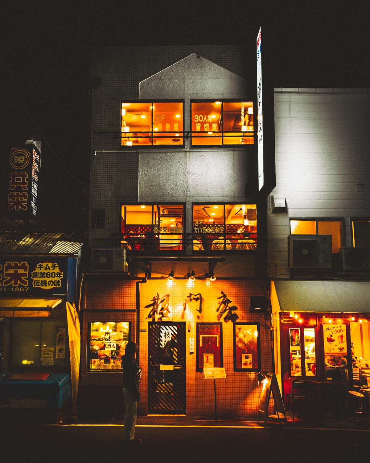 Chaleur nocturne d'une échoppe de quartier. La vie s'anime derrière les fenêtres éclairées.