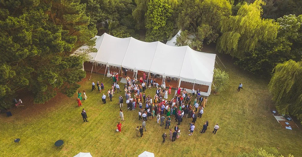Vue d'ensemble spectaculaire par drone d'un cocktail de mariage (vin d'honneur) sous un grand chapiteau blanc. Mise en valeur du lieu de réception et de l'ambiance champêtre.