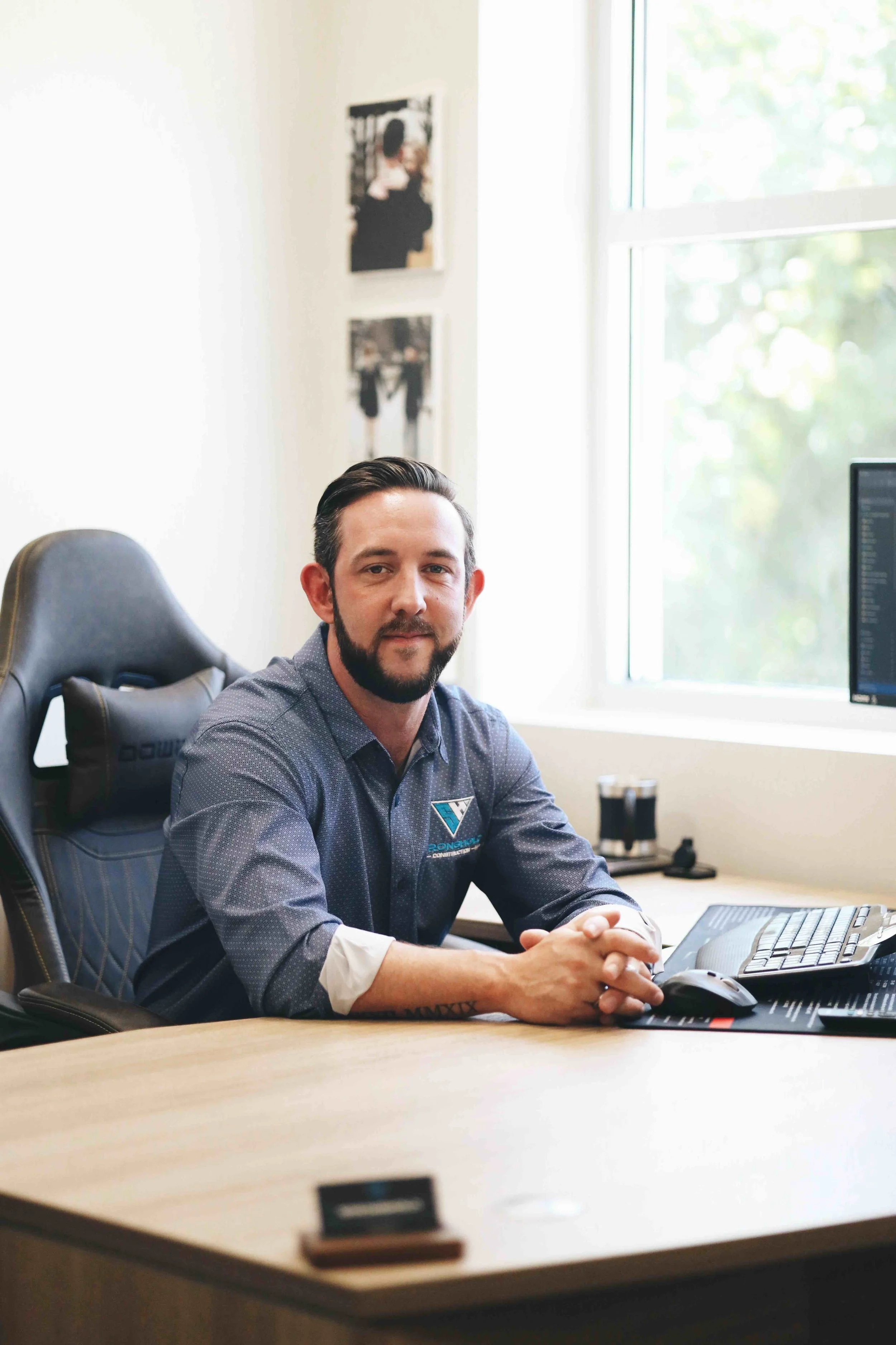 A man with dark hair and a beard sitting at a desk in an office, wearing a blue button-up shirt with a logo on it, in front of a computer with a large monitor, and a window behind him.