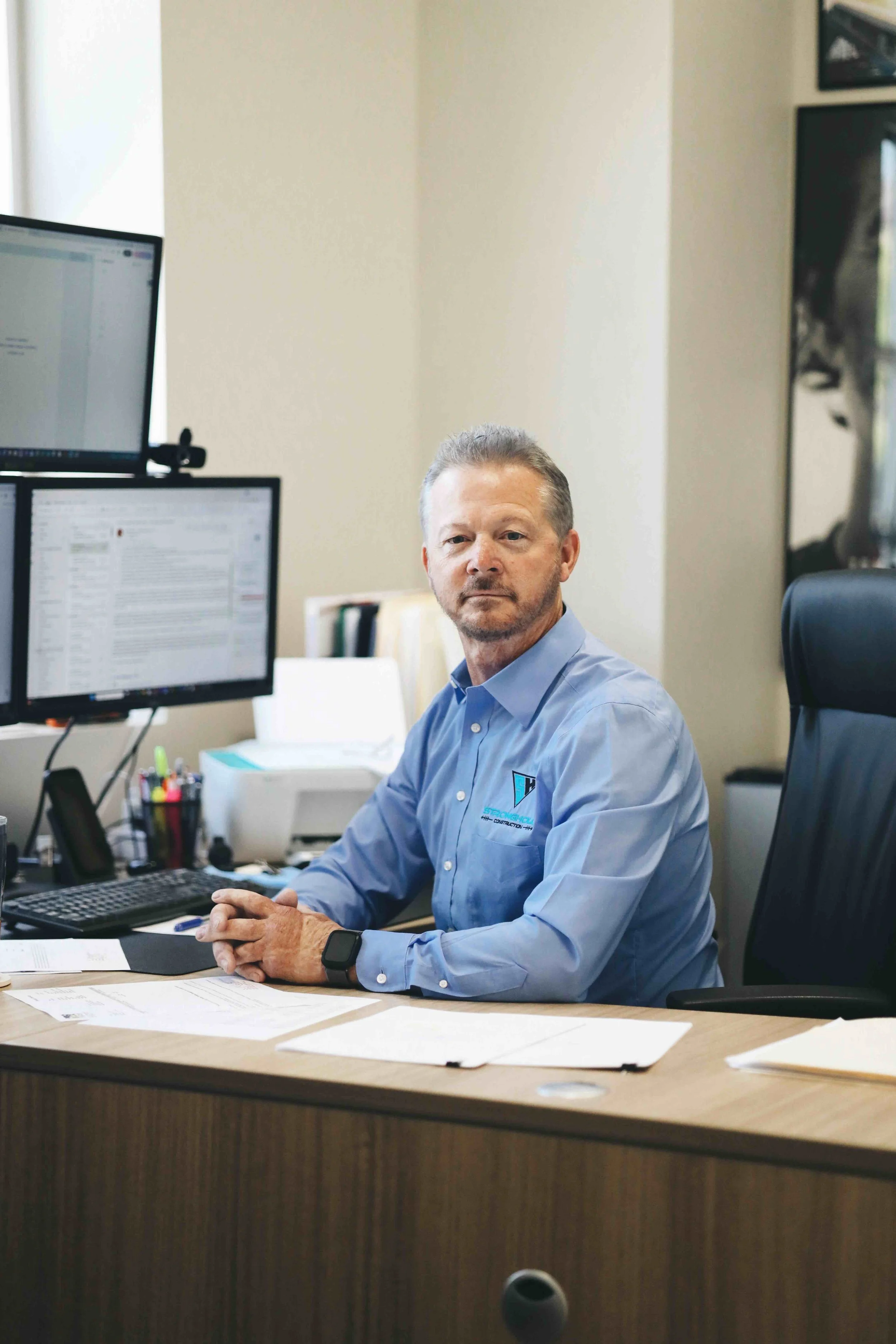 Man sitting at a desk with multiple monitors, papers, and office supplies in a professional office setting.