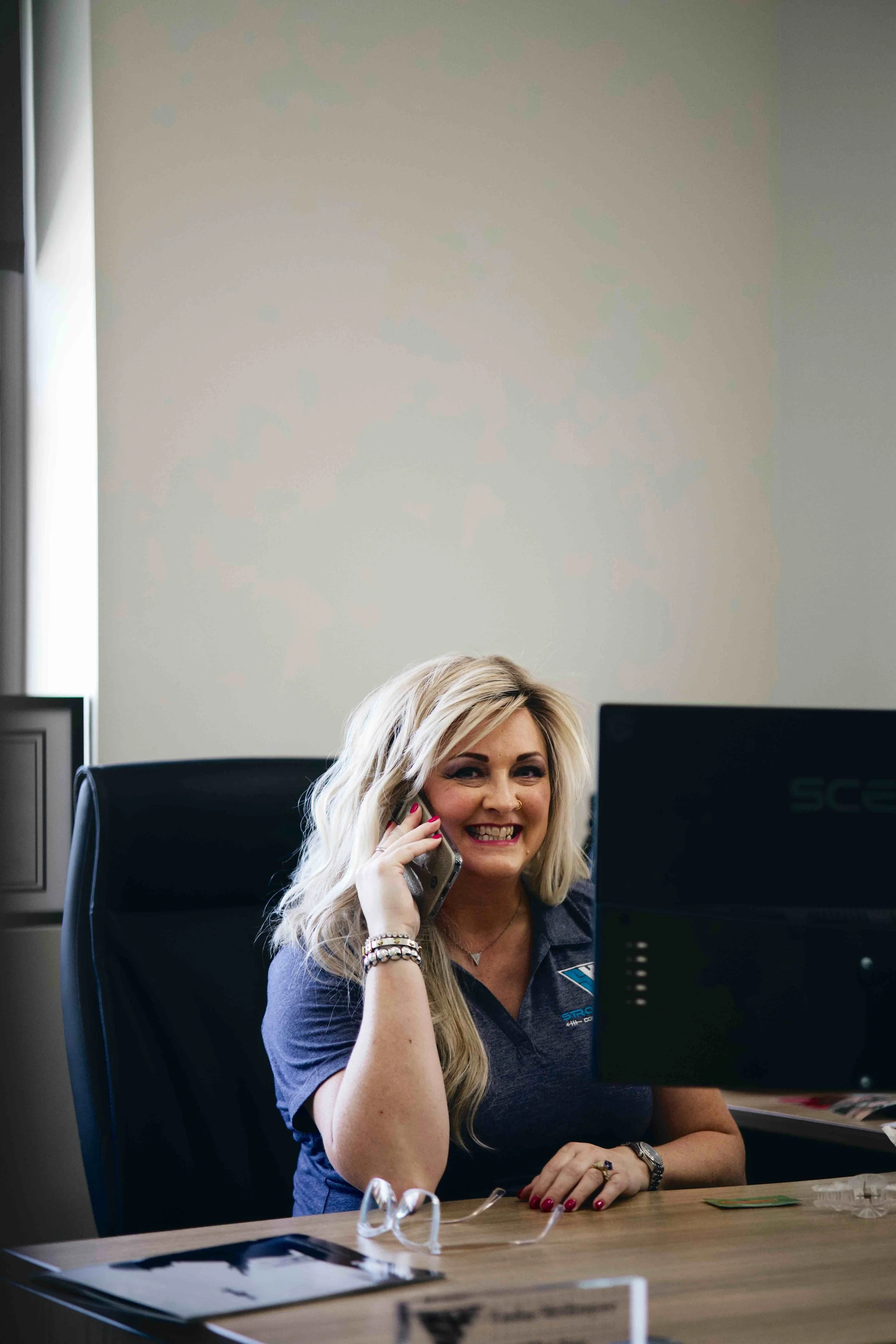 A woman with long blonde hair smiling while talking on a phone at her desk in an office.