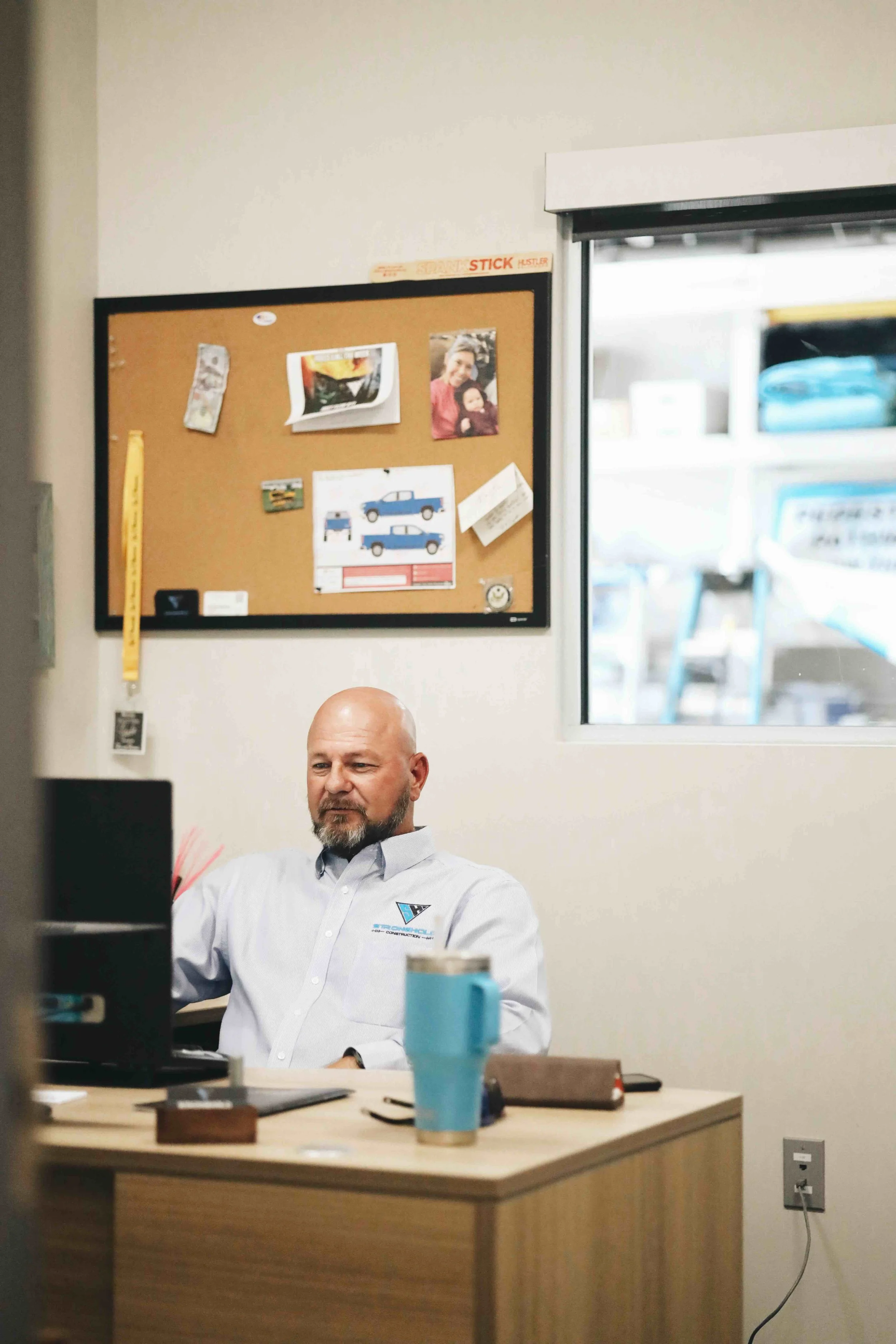 A man with a bald head and beard sitting at a desk in an office, working on a computer. The desk has a blue travel mug, a brown notebook, and some papers. Behind him, there is a cork board with various photos and notes, and a window showing a storage area with cars and shelves.