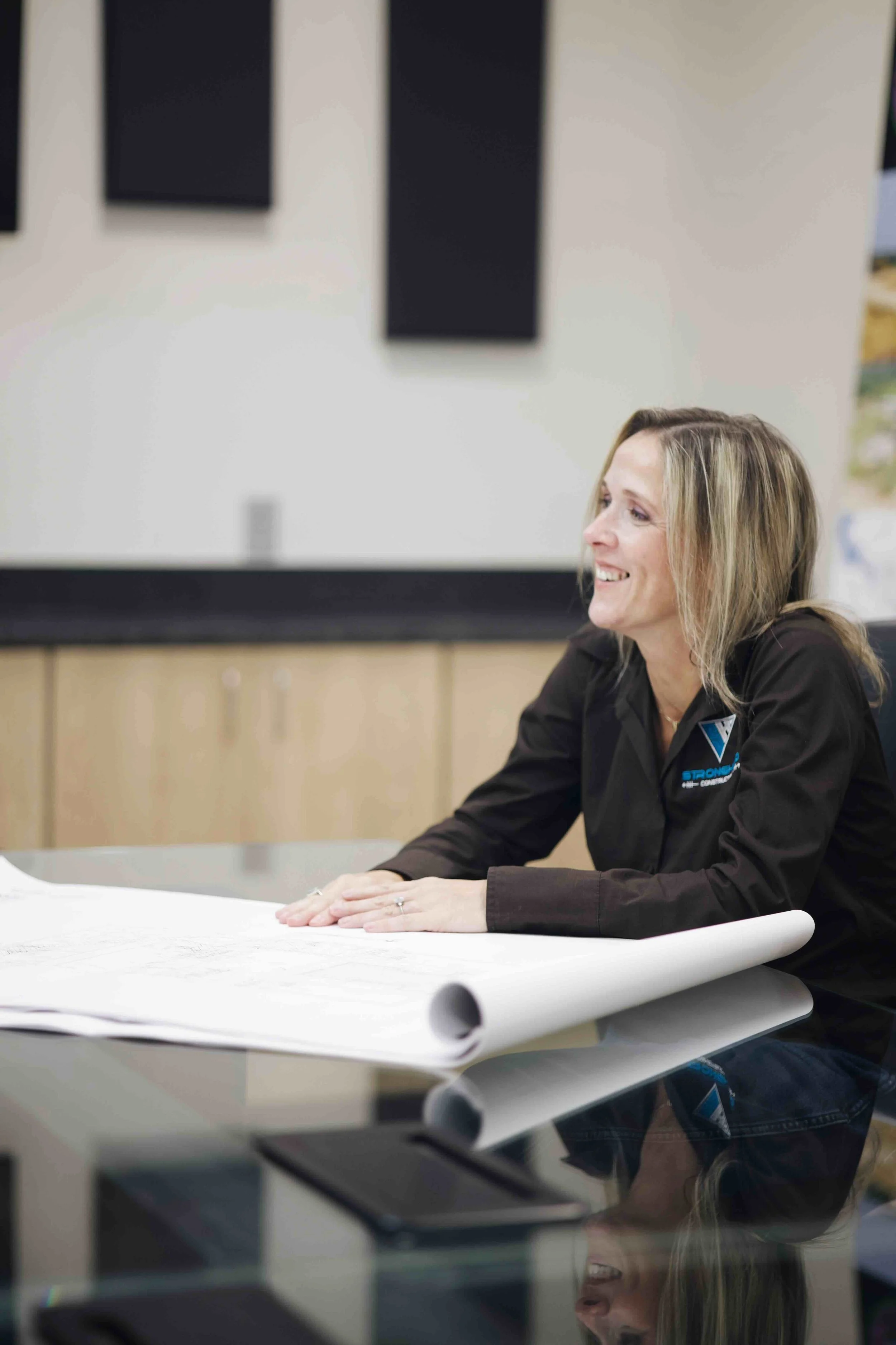 A woman smiling at a table with architectural blueprints, wearing a dark shirt with a logo.