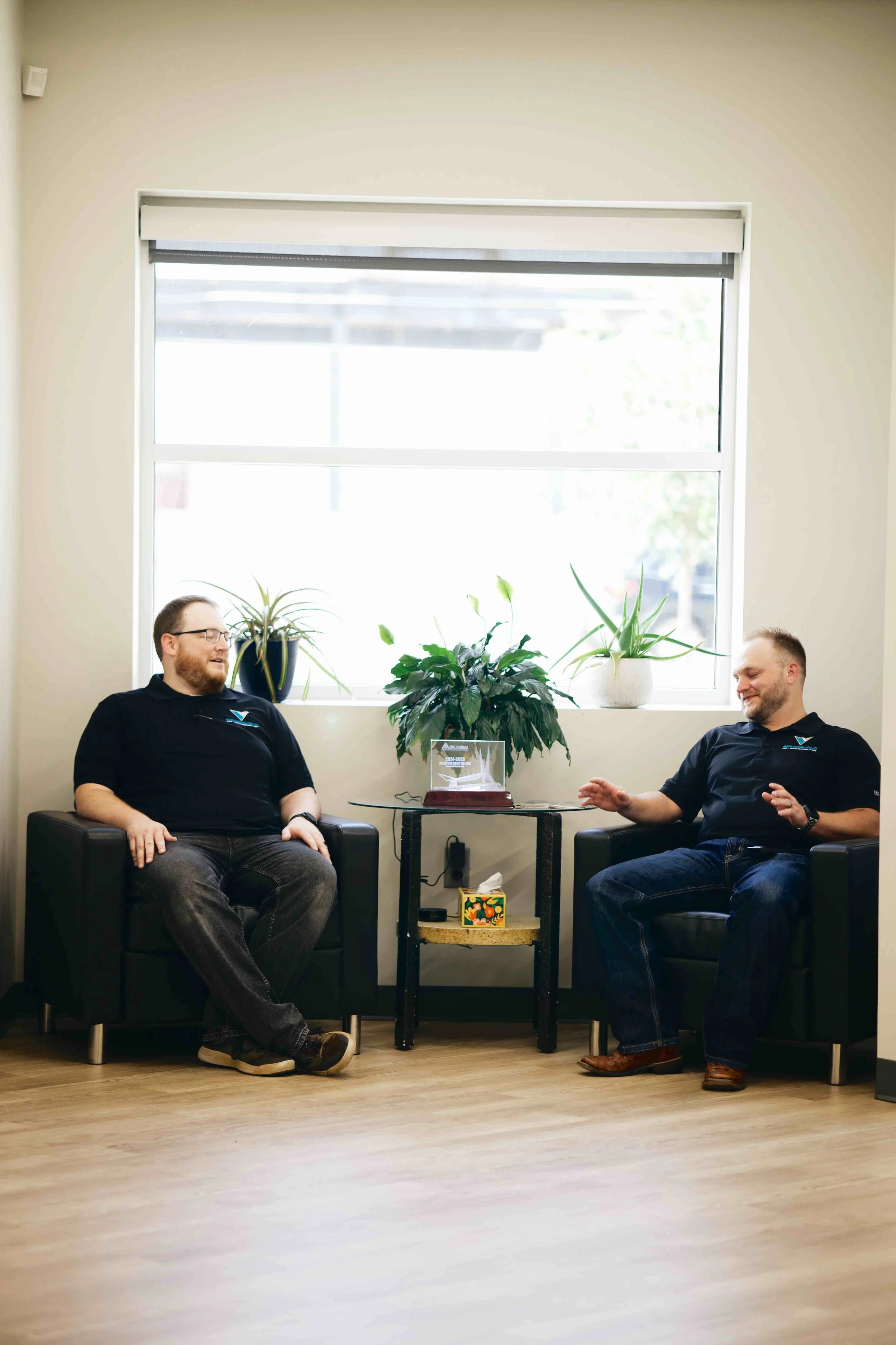 Two men sitting in black armchairs having a conversation in a room with a large window, potted plants, a small table with a decorative box and a framed item.