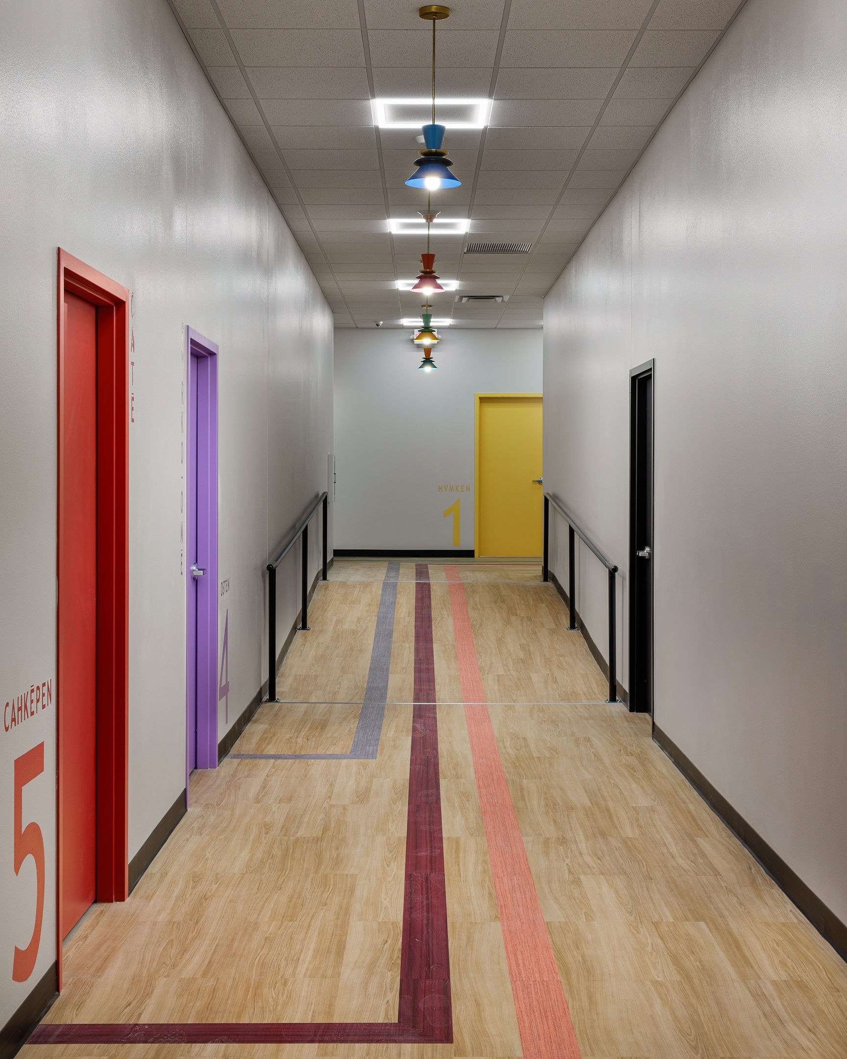 Ramped corridor with handrails and Mvskoke-numbered classroom doors resolving existing slab elevation changes at Barking Waters Head Start in Wewoka, Oklahoma