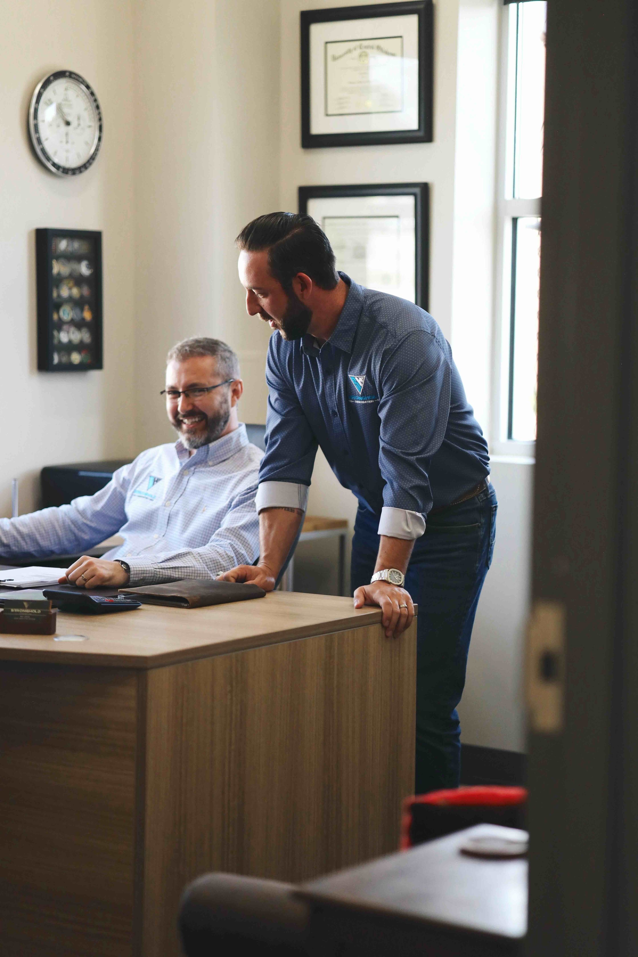 Two men in an office, one is sitting at a desk smiling, the other is standing leaning on the desk, engaged in conversation.
