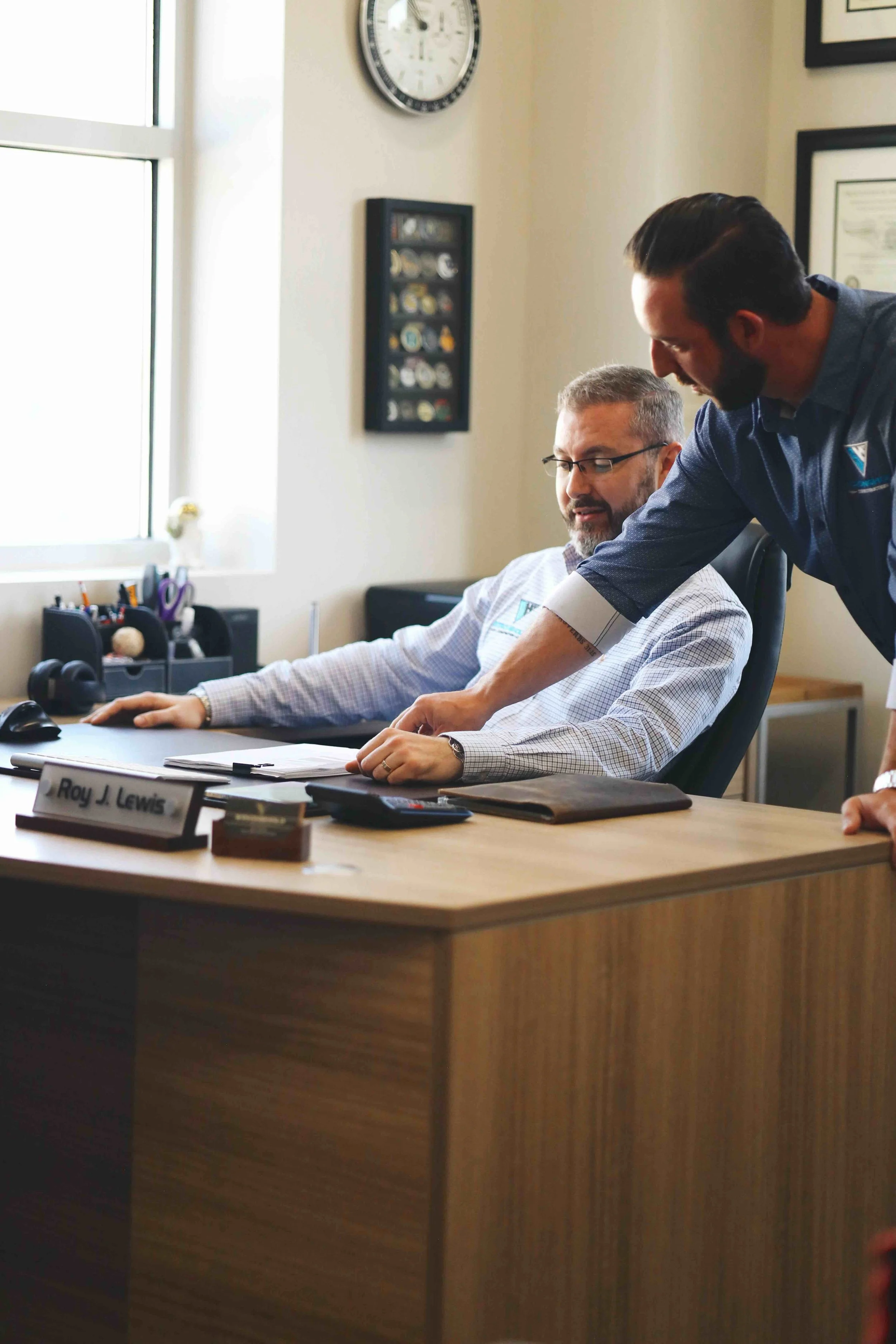 Two men are in an office; one is sitting at a desk with a nameplate that reads 'Roy J. Lewis' and the other is standing beside him, pointing at papers on the desk. The office has a window, a clock, framed certificates, and various office supplies.