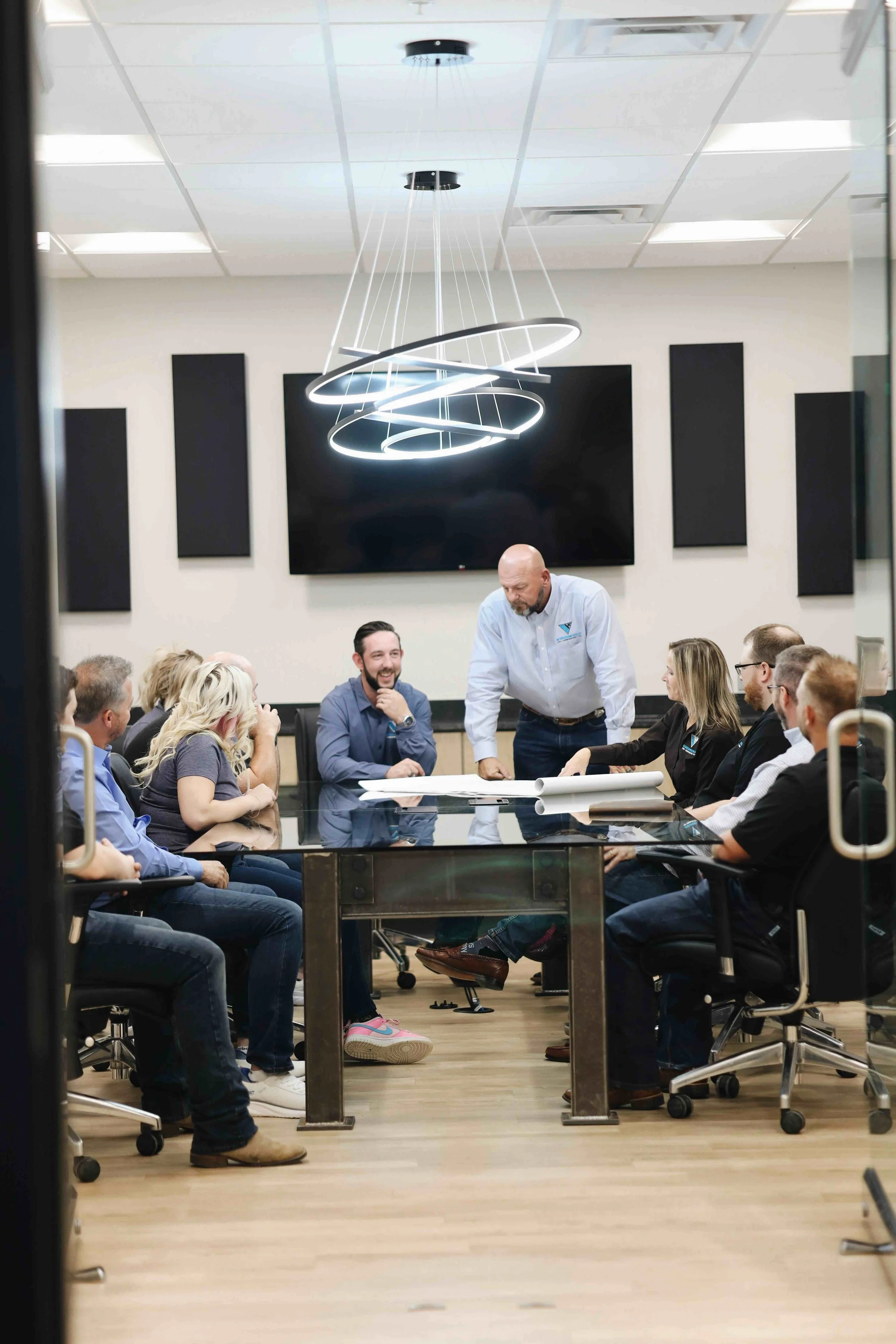 People in a business meeting or discussion, sitting around a glass conference table in a modern office, with a man standing and speaking.