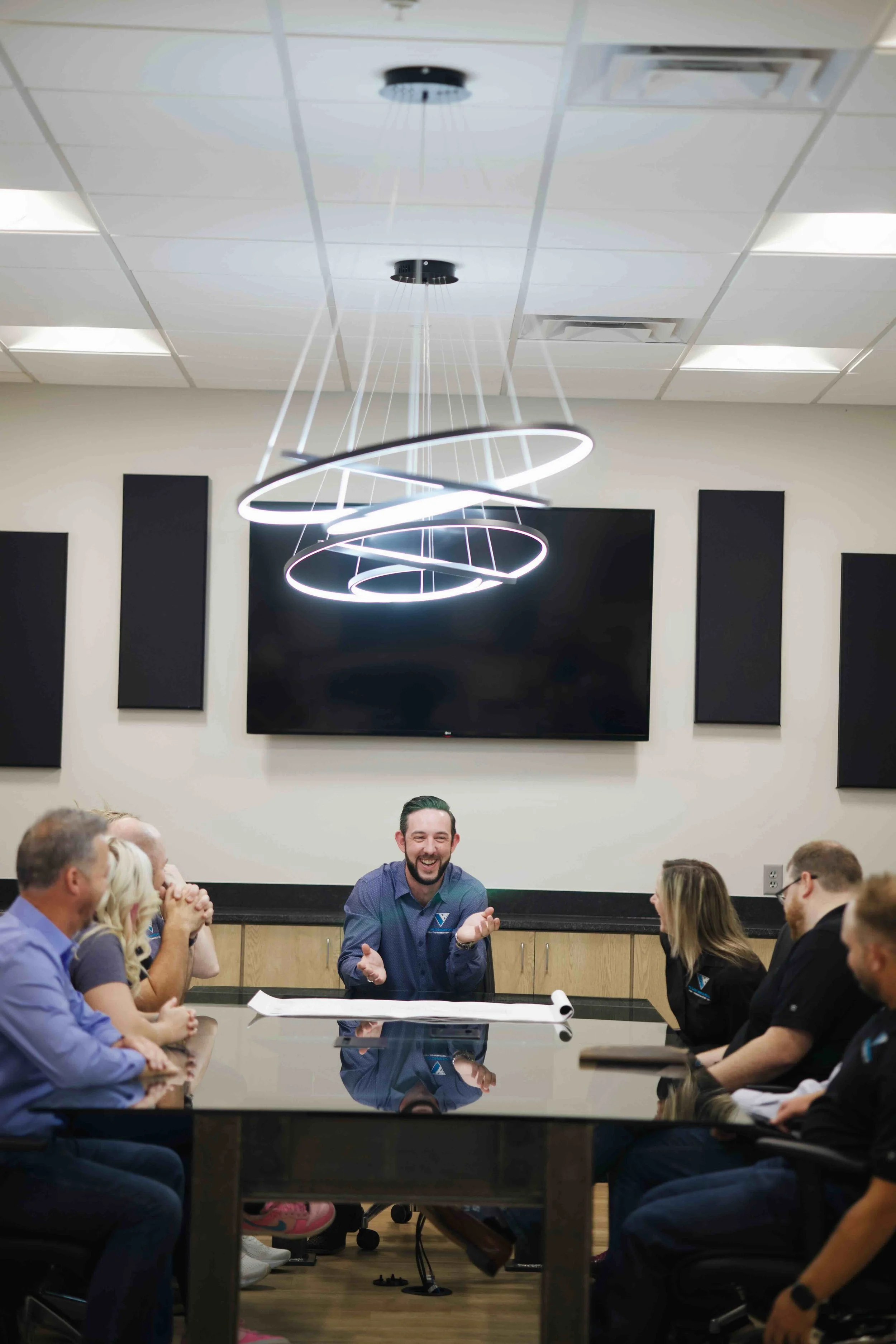 A group of people sitting around a conference table in a meeting room, listening to a man who is talking and smiling. The room has a large digital screen on the wall, and a modern ceiling light fixture.