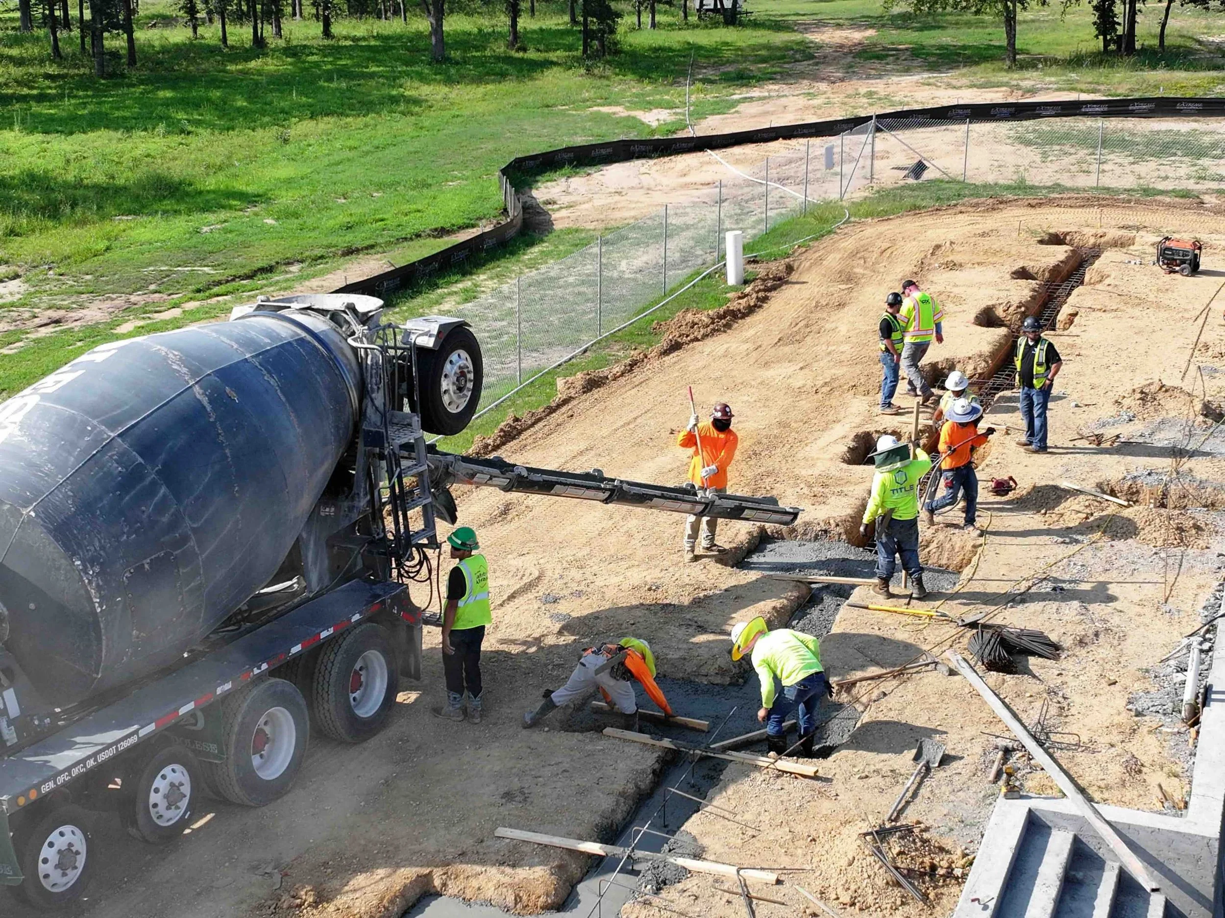 Construction workers are pouring and leveling concrete on a building foundation while a cement mixer truck is nearby on a dirt site surrounded by grassy areas and trees.
