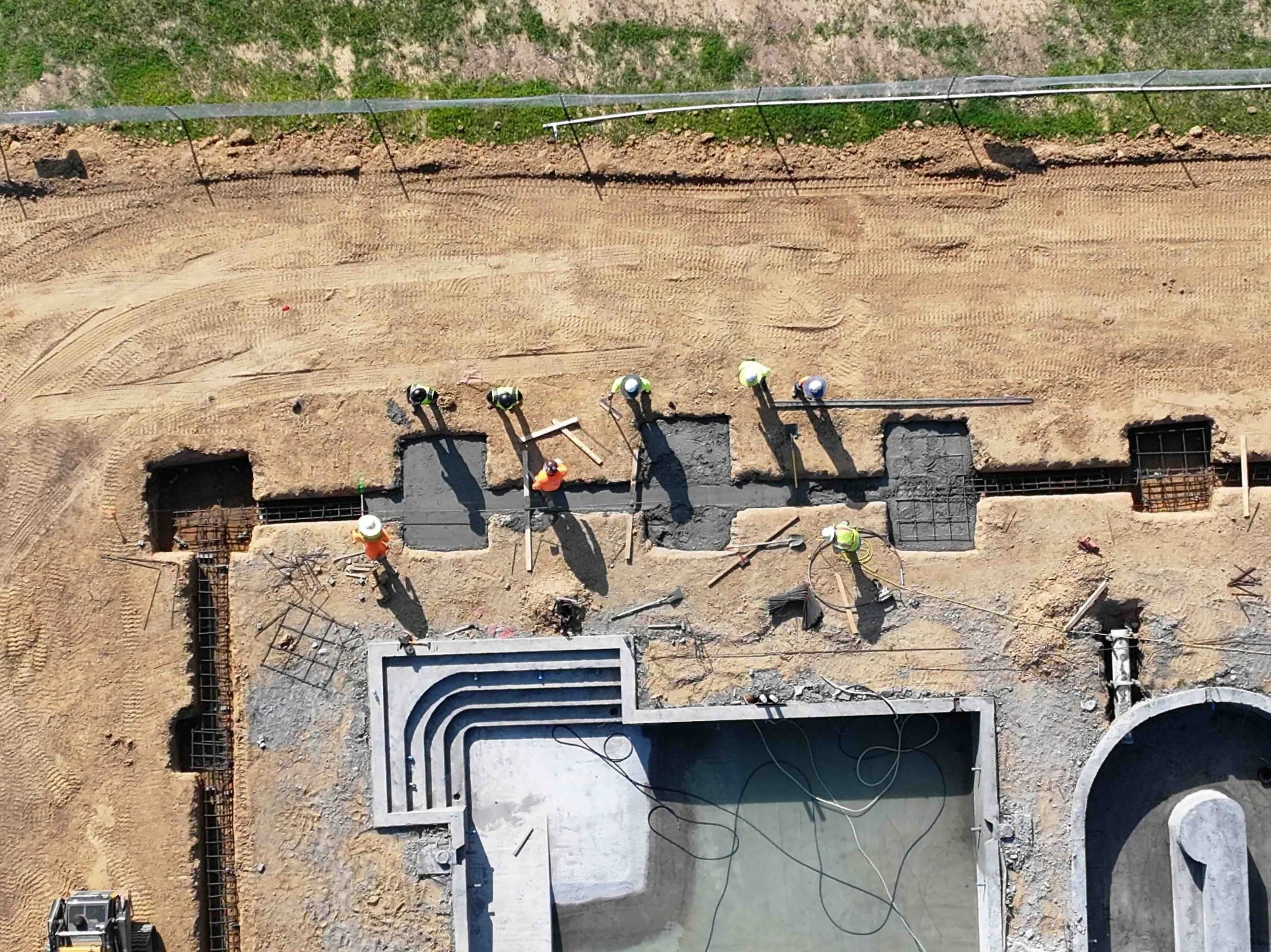 An aerial view of a construction site where workers are pouring concrete into long, narrow trenches. Several workers wearing safety helmets and vests are handling tools and guiding the concrete. The site is surrounded by dirt and some green grass.