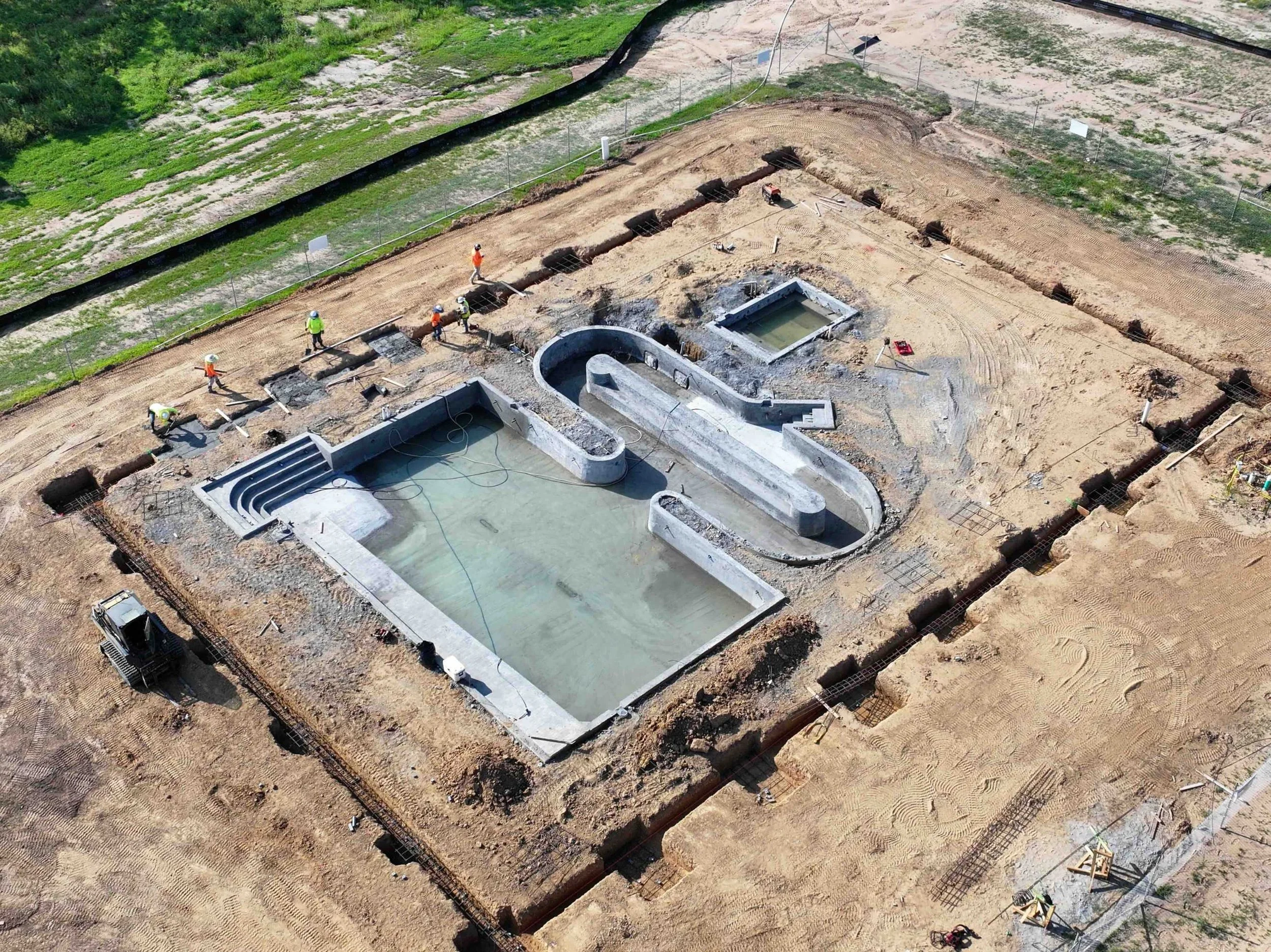 Under construction swimming pool with water, surrounded by dirt, with construction workers in safety gear working nearby.