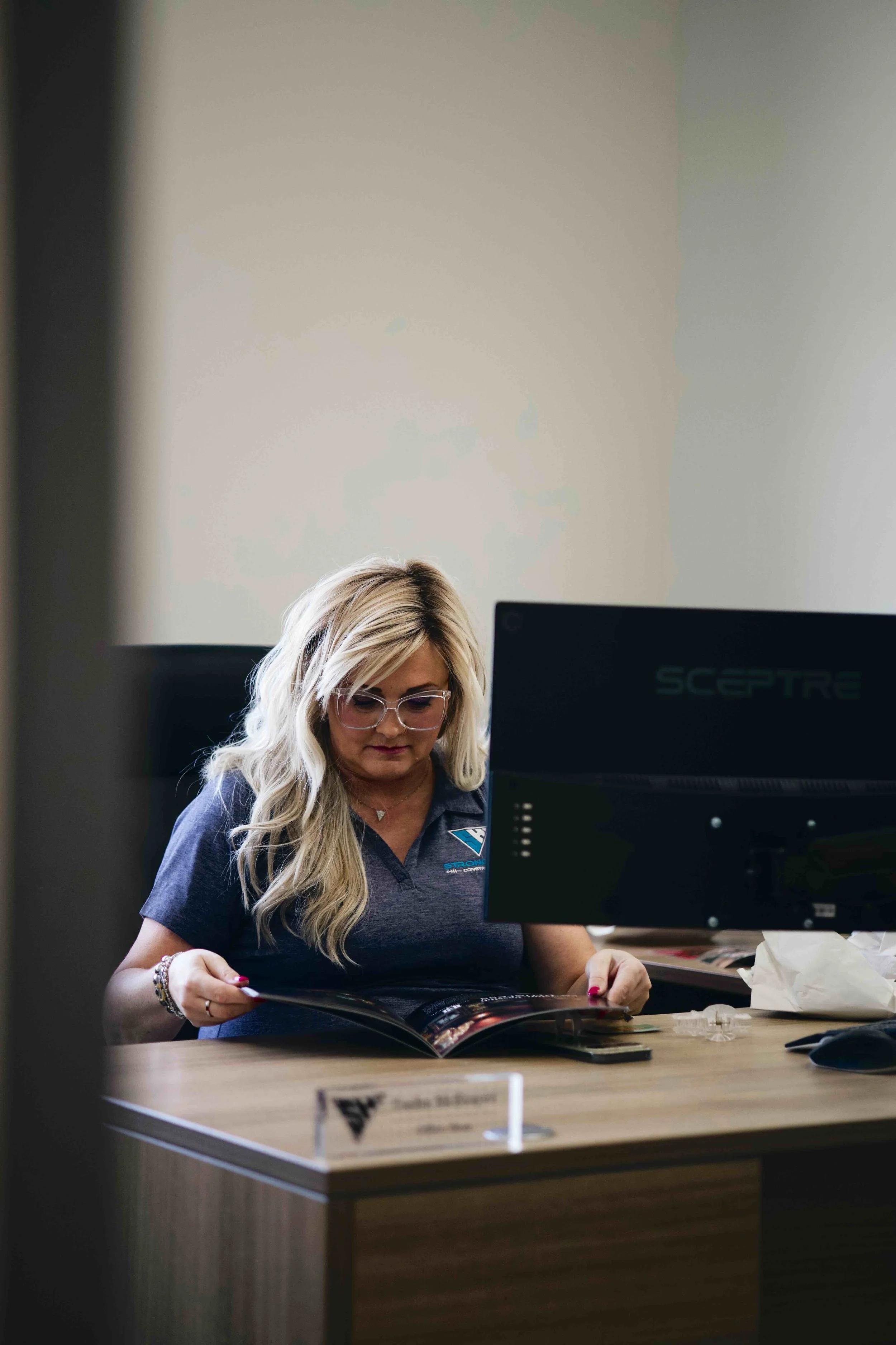 A woman with blonde hair and glasses sitting at a desk, reading a magazine, with a computer monitor in front of her.