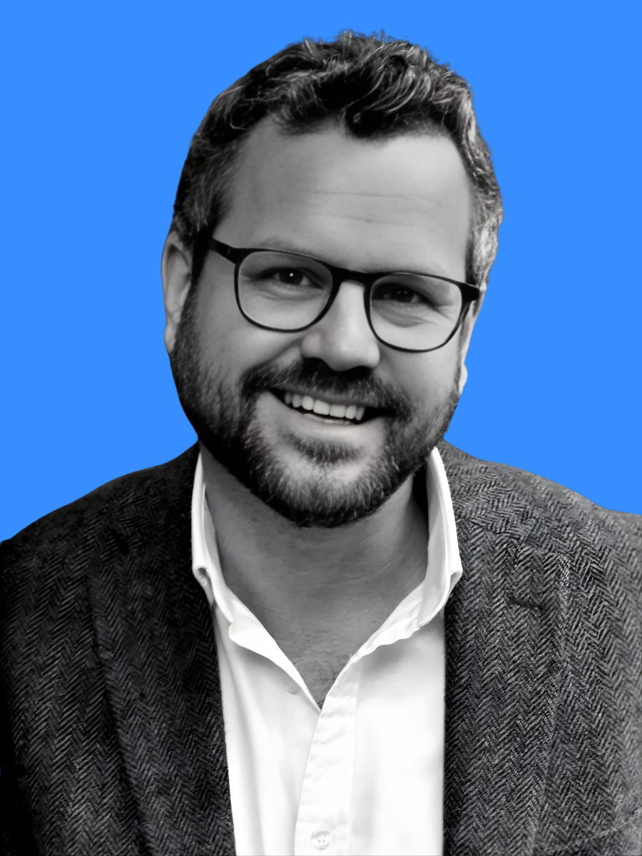 A smiling man with glasses, a beard, and short, wavy hair posing against a solid blue background. He is wearing a blazer over a white shirt.