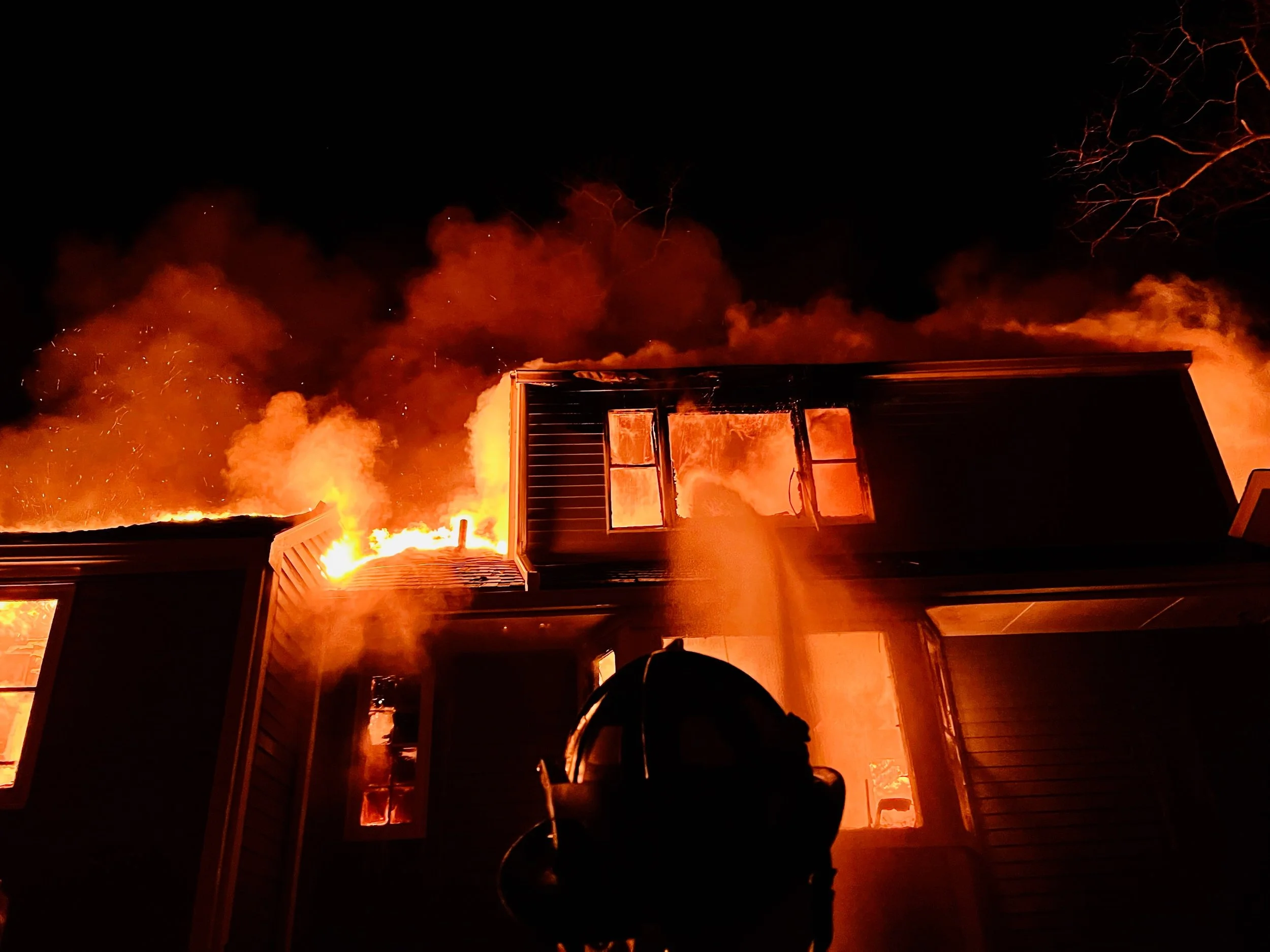 A house on fire at night with flames and thick smoke billowing from the roof, with firefighter equipment visible in the foreground.
