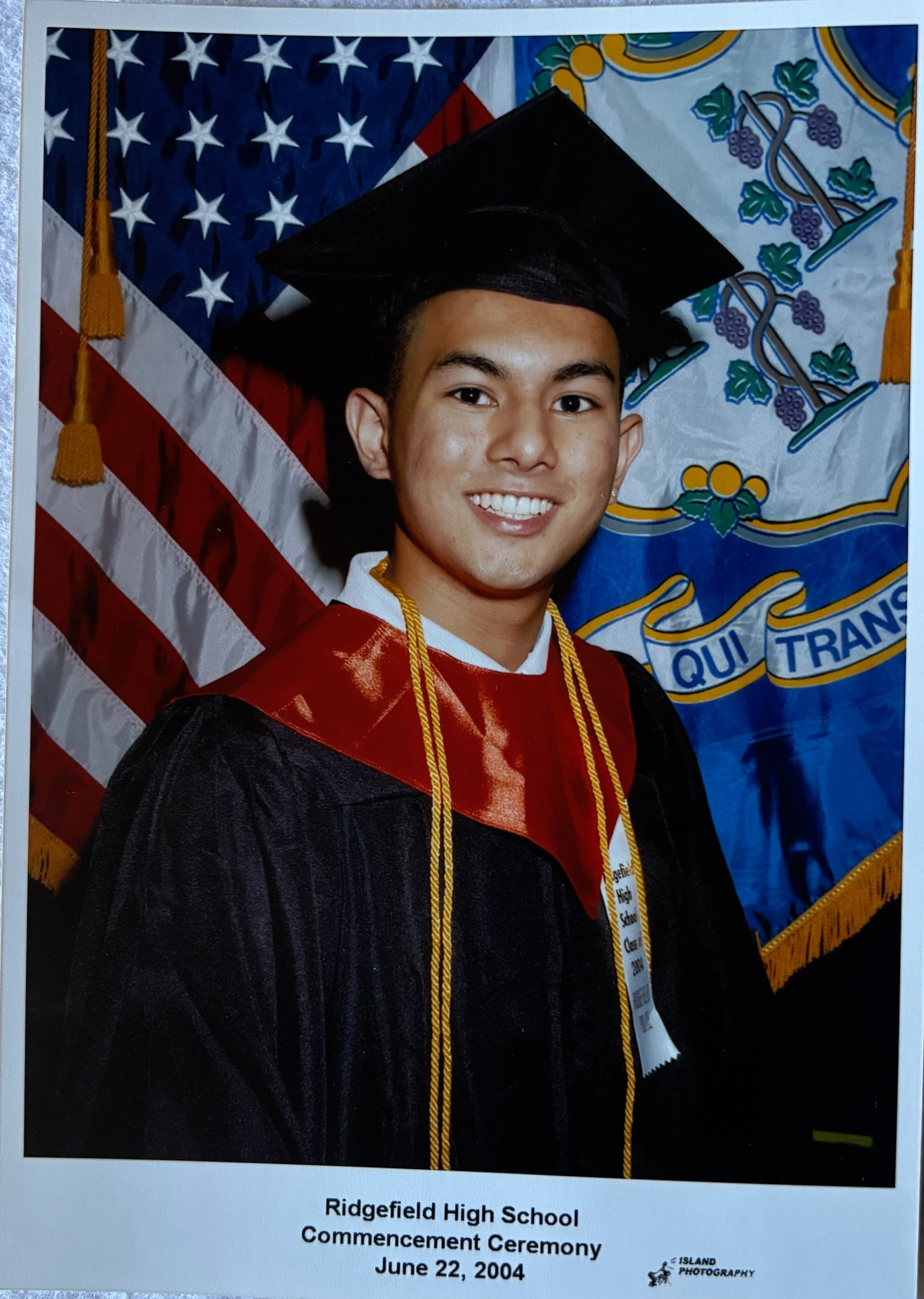 A young man wearing a graduation cap and gown with honor cords, smiling, at Ridgefield High School's commencement ceremony on June 22, 2004, with American and school flags in the background.