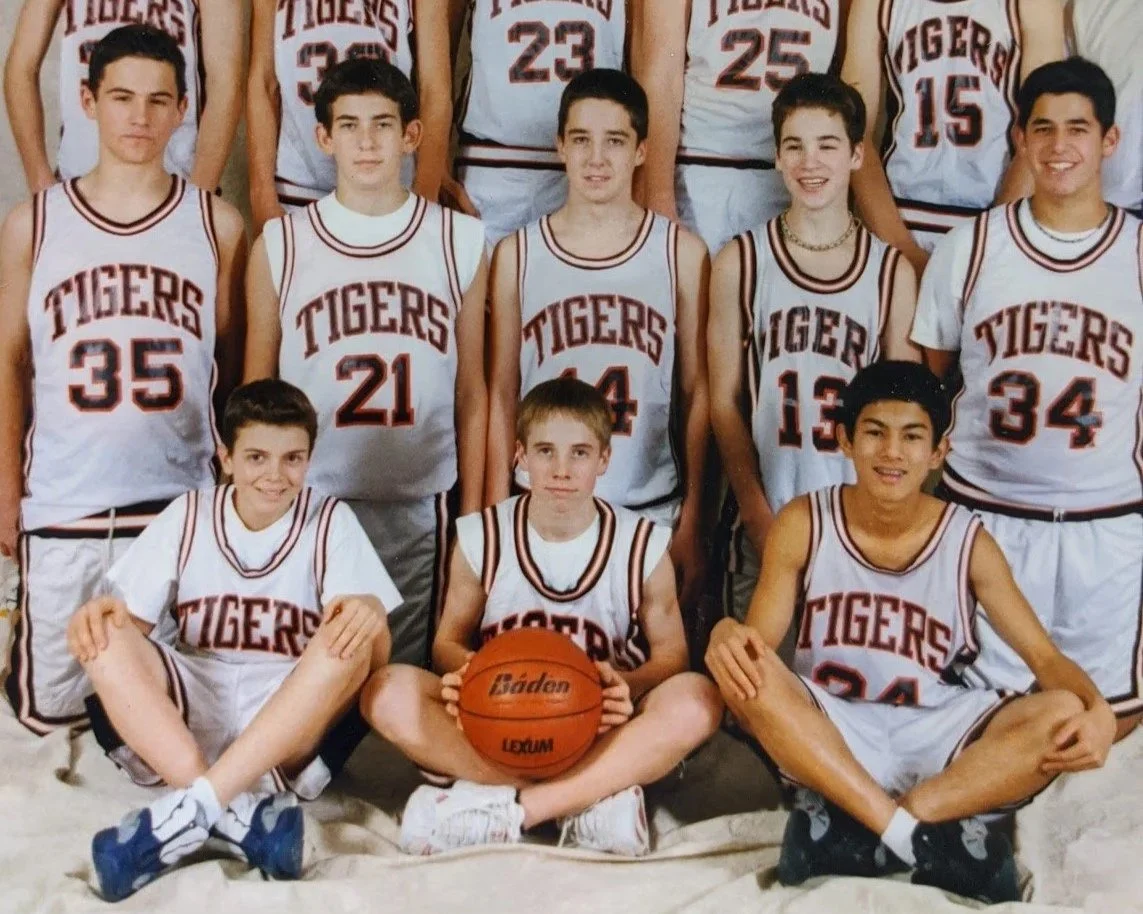 A group of young boys in basketball uniforms posing for a team photo, with some sitting Cross-legged in front and others standing behind. They are wearing white jerseys with red and black accents that say 'TIGERS' and have various numbers. One of the boys in the front is holding a basketball.
