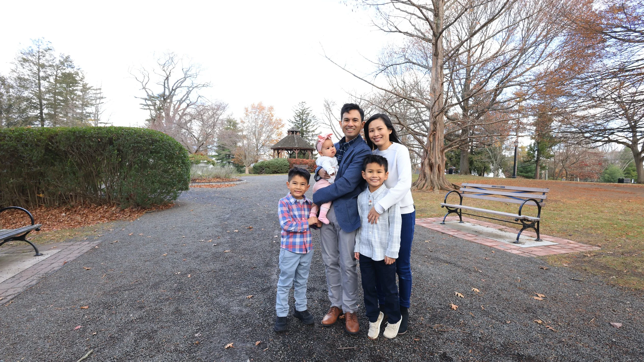 A family of five standing on a gravel path in a park during fall, with leafless trees and benches around them.